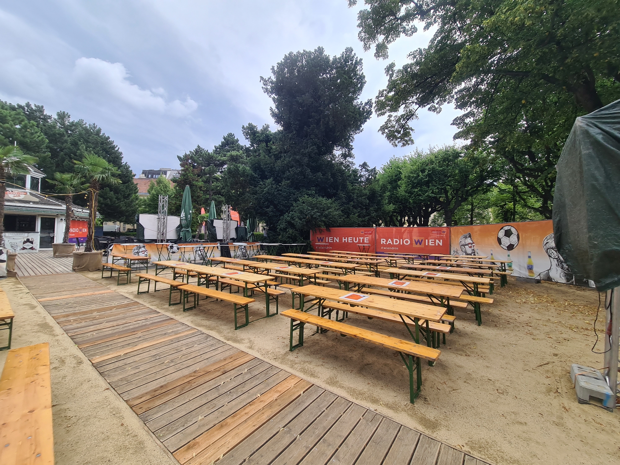 The image depicts an outdoor seating area with numerous picnic tables and benches arranged on a sandy ground. The tables are made of light-colored wood with green metal legs. Several tables have orange square markers on their surfaces. In the background, there's a large orange banner with the text "WIEN HEUTE RADIO WIEN" and images of people and a soccer ball. Green umbrellas are folded and placed behind the tables. Trees form a backdrop, and a cloudy sky is visible above. A wooden walkway runs through the foreground. The scene appears to be set up for an event, possibly a public viewing or gathering, in Vienna, Austria, suggested by the banner text. There are no people present in the scene.