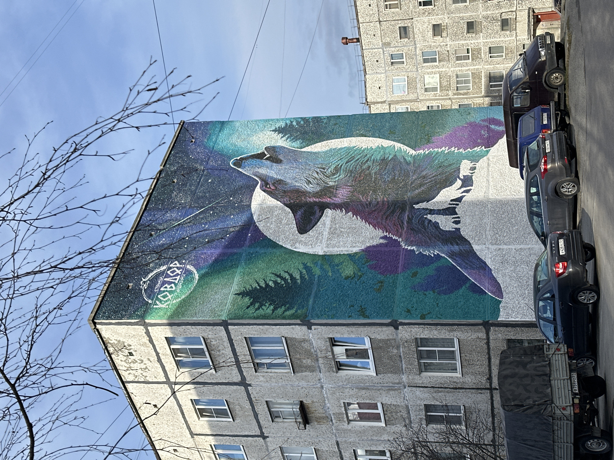 The image depicts the side of a multi-story building in an urban setting. A large mural of a howling wolf, set against a full moon and a backdrop resembling the aurora borealis, adorns the side of the building. Text in a foreign language is visible in a circular emblem on the mural. The building itself appears to be constructed from concrete and features a grid of windows. Several vehicles are parked along the street adjacent to the building, including a truck with a covered bed and several cars in shades of gray, blue, and black. A bare tree is visible in the foreground. The sky is light blue with some sparse clouds.
