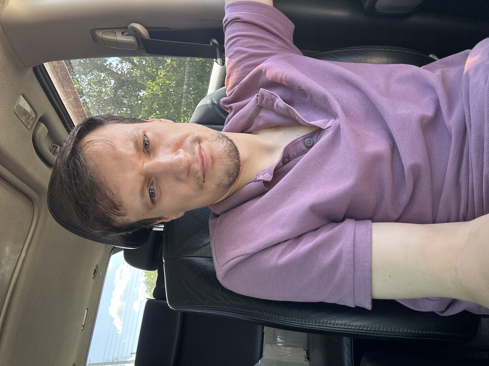 A man is sitting in the driver's seat of a car. He is wearing a purple polo shirt and has short brown hair. The interior of the car is visible, including black leather seats, a sunroof, and a seatbelt. Through the windows, trees and a blue sky with clouds can be seen. The image was taken outdoors in the city of Makiv, Ukraine.