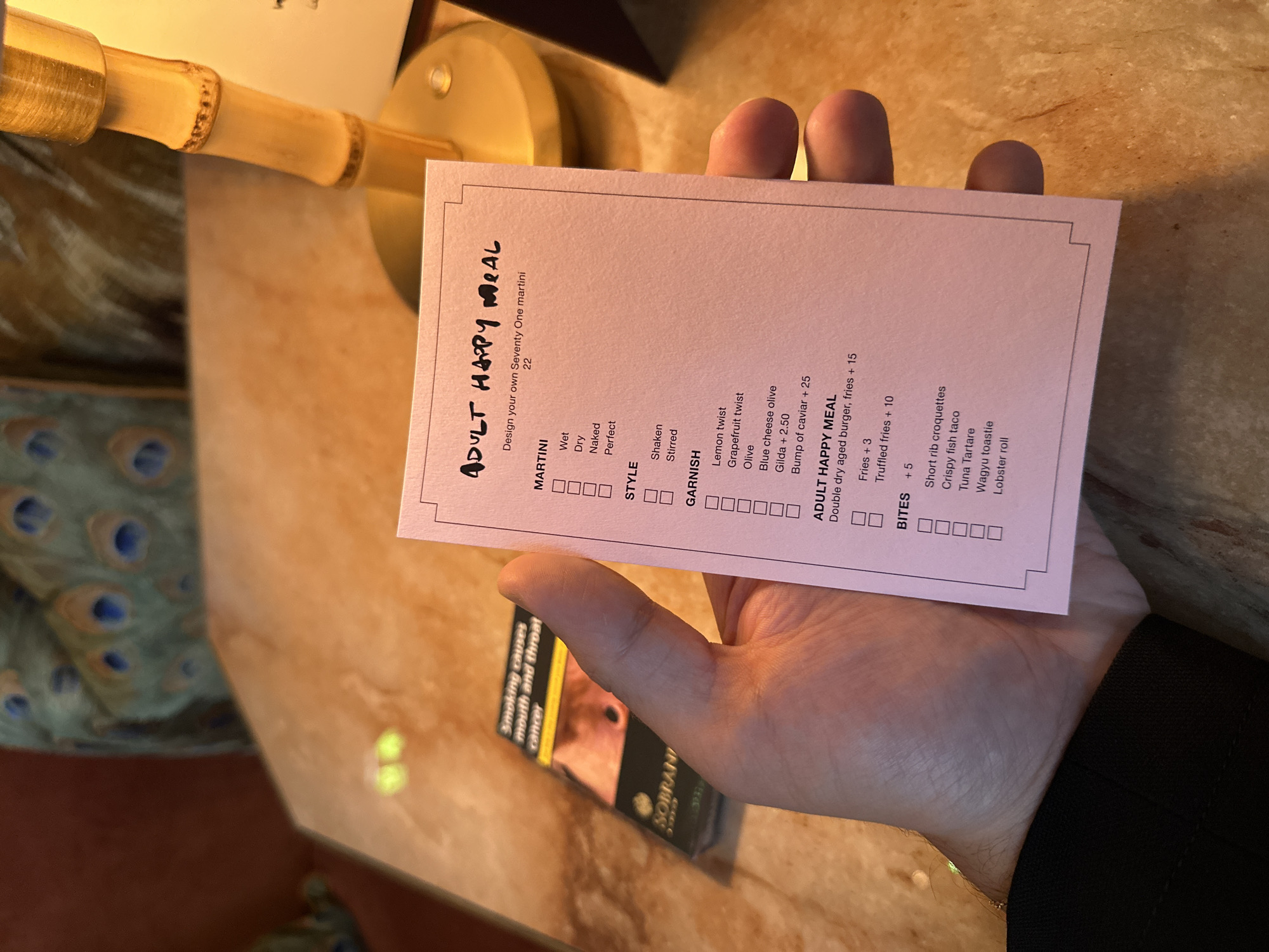 A close-up, eye-level shot captures a hand holding a pink menu with the title "ADULT HAPPY MEAL" printed in handwritten black marker at the top. Below the title, it reads "Design your own Seventy One martini 22". The menu presents options for "MARTINI" (Wet, Dry, Naked, Perfect), "STYLE" (Shaken, Stirred), "GARNISH" (Lemon twist, Grapefruit twist, Olive, Blue cheese olive, Gilda + 2.50, Bump of caviar + 25), and "ADULT HAPPY MEAL" (Double dry aged burger, fries + 15). It also lists "BITES" with options like "+ 5", "Short rib croquettes", "Crispy fish taco", "Tuna Tartare", "Wagyu toastie", and "Lobster roll."
In the background, a pack of Sobranie cigarettes with a prominent warning label about smoking is visible. A part of a lamp with a bamboo-style base and a golden shade is also in the frame. The setting appears to be a table in a dimly lit, upscale environment, possibly a bar or restaurant, judging by the decor which includes ornate peacock feather patterns on the wall and a textured surface of the table. The image is taken indoors. The overall scene is casual, suggesting a person is about to order or has just received their menu.