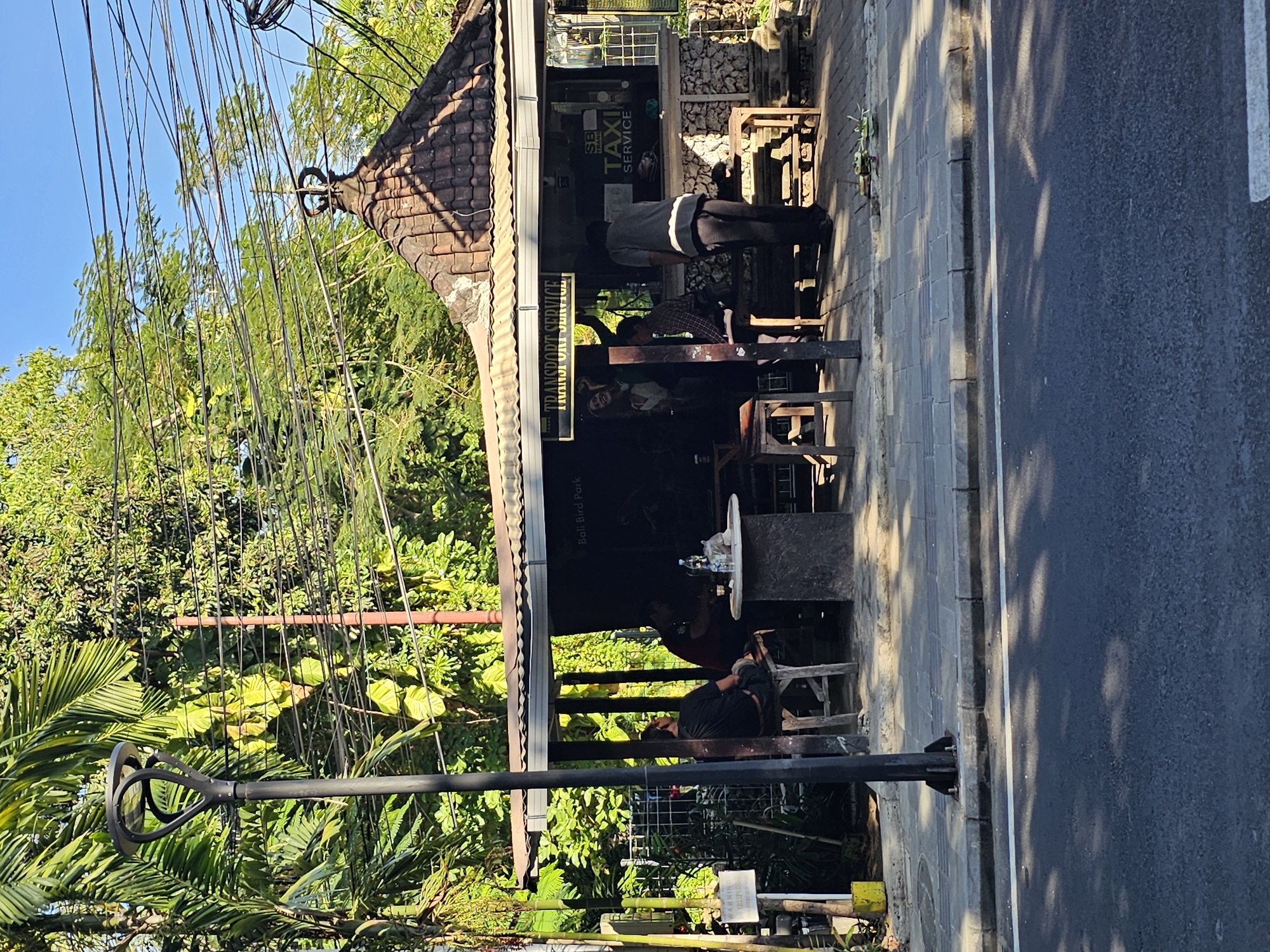 The image depicts an outdoor scene in Denpasar, Indonesia. In the foreground, a paved road with a white line marking the lane is visible. Adjacent to the road is a concrete sidewalk with a gray tiled surface and a curb. A black street lamp stands on the sidewalk.
Behind the sidewalk is a covered open-air structure, possibly a small shop or service area. The roof is made of terracotta tiles, and the structure has an open front. A sign above the entrance reads "TRANSPORT SERVICE". Inside and around the structure, several people are present. Two men are seated at a table in the shade, with bottles and what appears to be food on the table. Another person stands near the counter, and others are partially visible inside. A sign for "SB TRANS TAXI SERVICE" is visible on the structure.
The background is filled with lush green foliage, including palm trees and other dense vegetation, suggesting a tropical environment. Power lines crisscross the sky. The overall impression is a roadside scene in a tropical urban setting, likely a service point or rest stop.