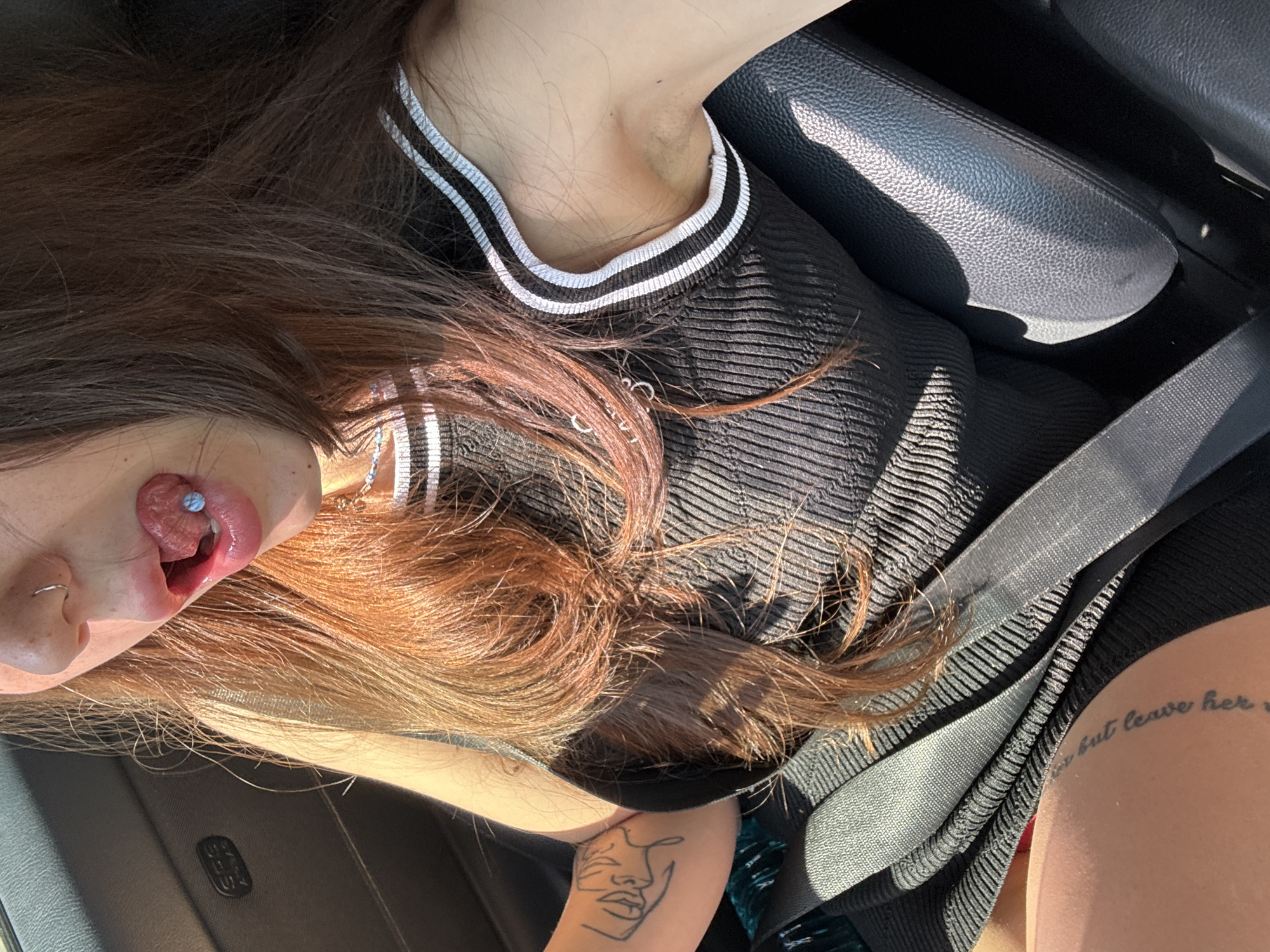 A woman is seen sitting in the passenger seat of a car, possibly in Sofia, Bulgaria. The seat belt is fastened across her torso. She is wearing a black ribbed sleeveless top with white stripes on the collar. Her tongue, pierced with a blue stud, is partially extended from her mouth. Her hair is long and auburn. A tattoo of a profile can be seen on her left arm, and a script tattoo is visible on her right thigh. The interior of the car is dark, and sunlight casts shadows on the seats and the woman.