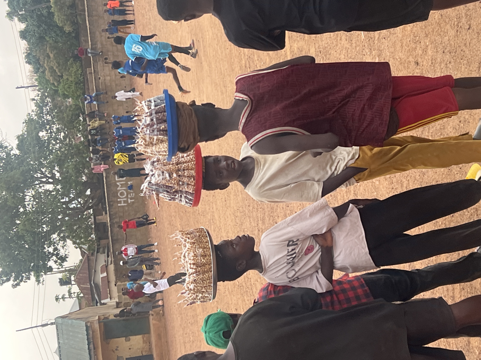Three individuals balance large platters of snacks atop their heads. In the background, a soccer field with players and spectators is visible. The location is Jos, Nigeria.