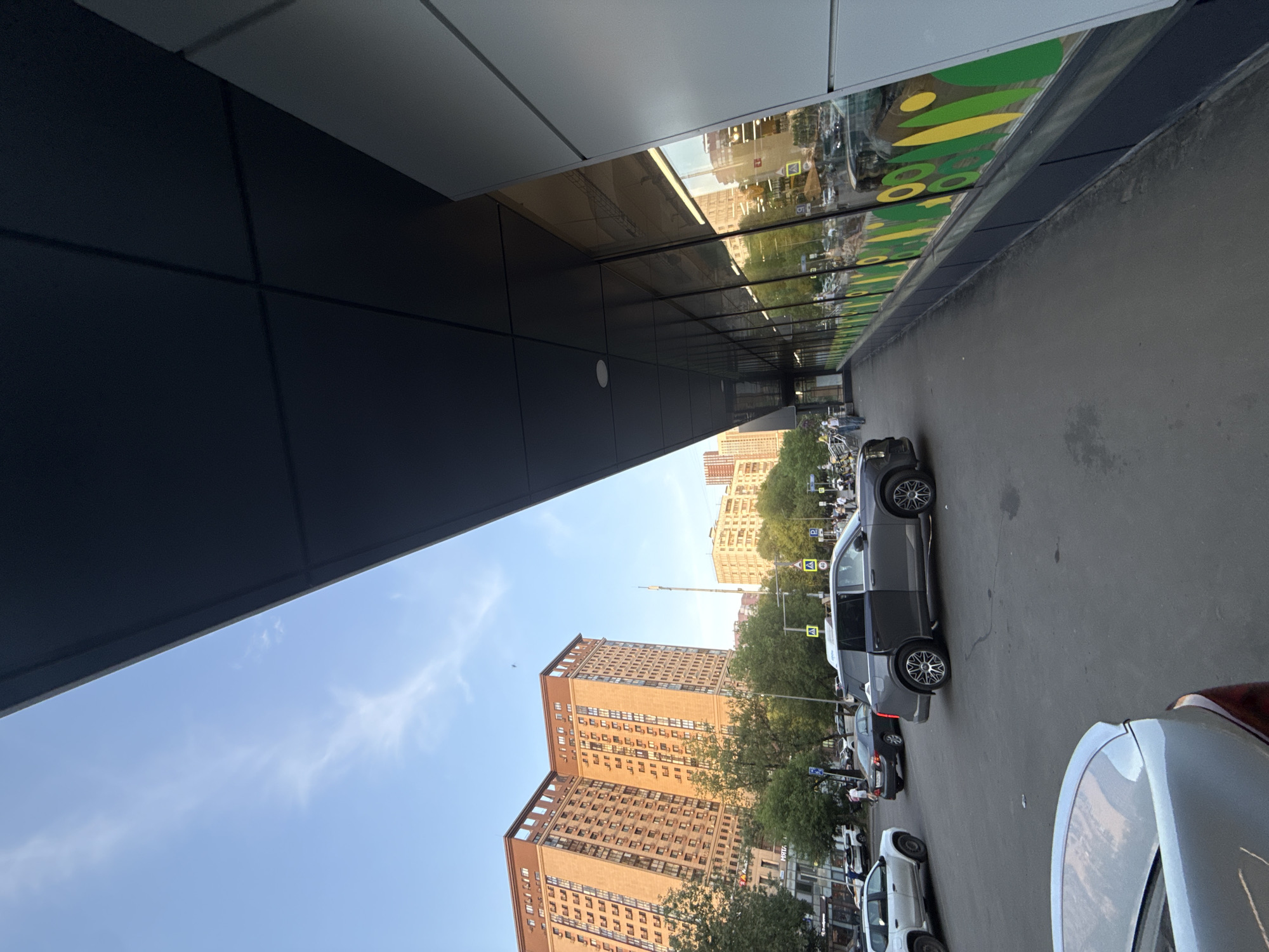 The image captures a street scene in Moscow, Russia, from a perspective under the overhang of a modern building. The sky is light blue with some clouds visible. In the background are large, tan-colored buildings. Green trees line the street. Parked cars are visible along the curb, and there are people walking on the sidewalk. There are also road signs with yellow triangles. The modern building's facade is gray and reflects the street scene in its windows.