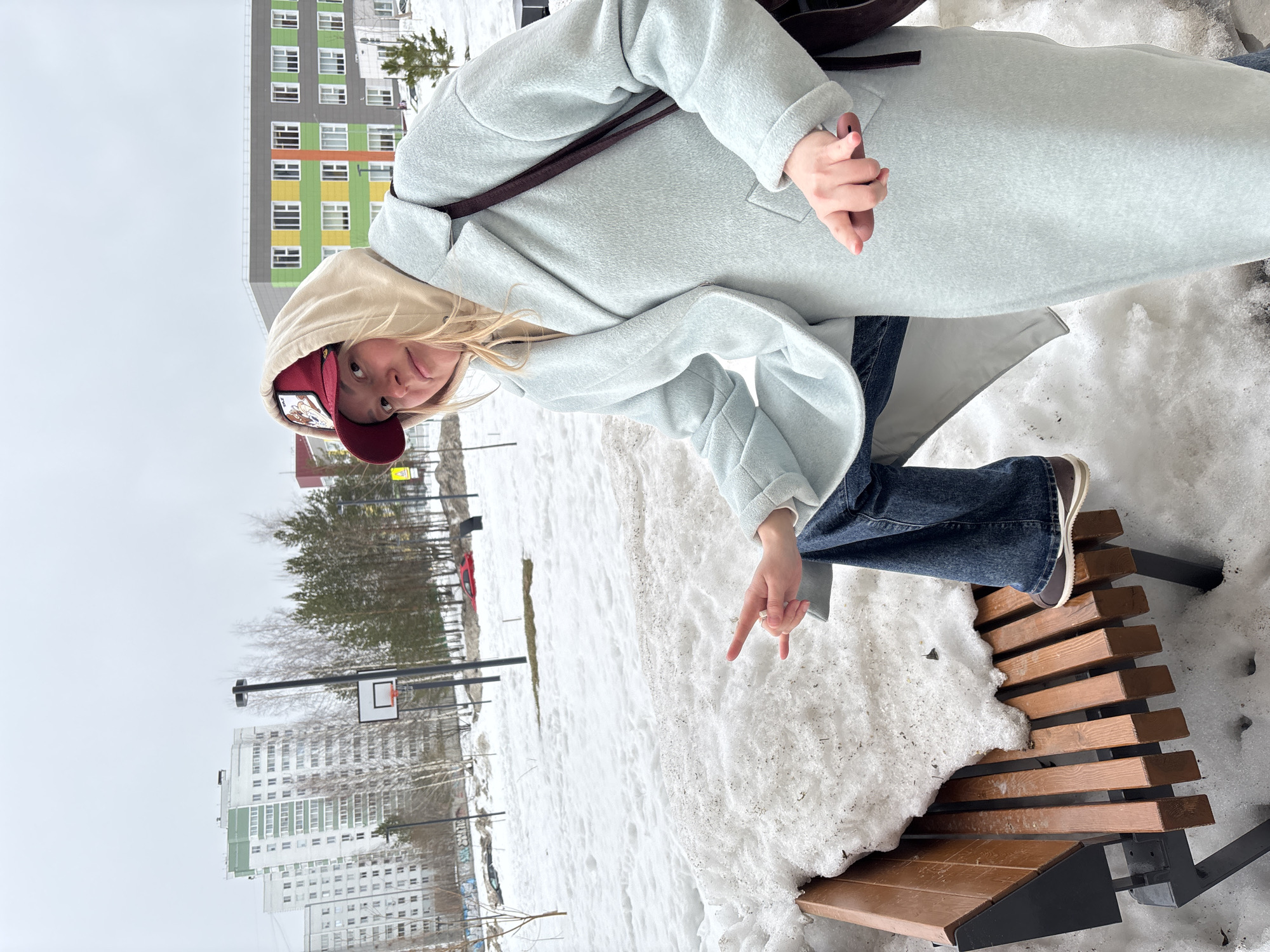 The image depicts an urban outdoor scene in Izhevsk, Russia, under an overcast sky, characterized by significant snow cover, indicating late winter or early spring conditions. A single female individual, appearing to be a young adult, is positioned centrally, facing the camera. She wears a maroon baseball cap featuring an animal emblem, a beige hooded sweatshirt, a light blue long coat, and dark blue jeans. Her left foot is resting on a snow-covered wooden slatted bench, which exhibits both clean wood and dirty snow accumulation. Her left hand is raised, with the index and pinky fingers extended in a "rock on" gesture, and a silver ring is visible on her left ring finger. Her right hand is held lower, grasping a dark-colored object, likely a smartphone. The background features multiple multi-story apartment buildings; one is predominantly white with green-paneled sections, and another is modern and colorful, displaying panels of green, yellow, orange, and grey. The environment includes bare deciduous trees, evergreen pines, a basketball hoop with a white backboard on a black pole, and streetlights. A red vehicle is partially visible behind a snowdrift in the midground, and a yellow triangular sign is discernible on the far right.