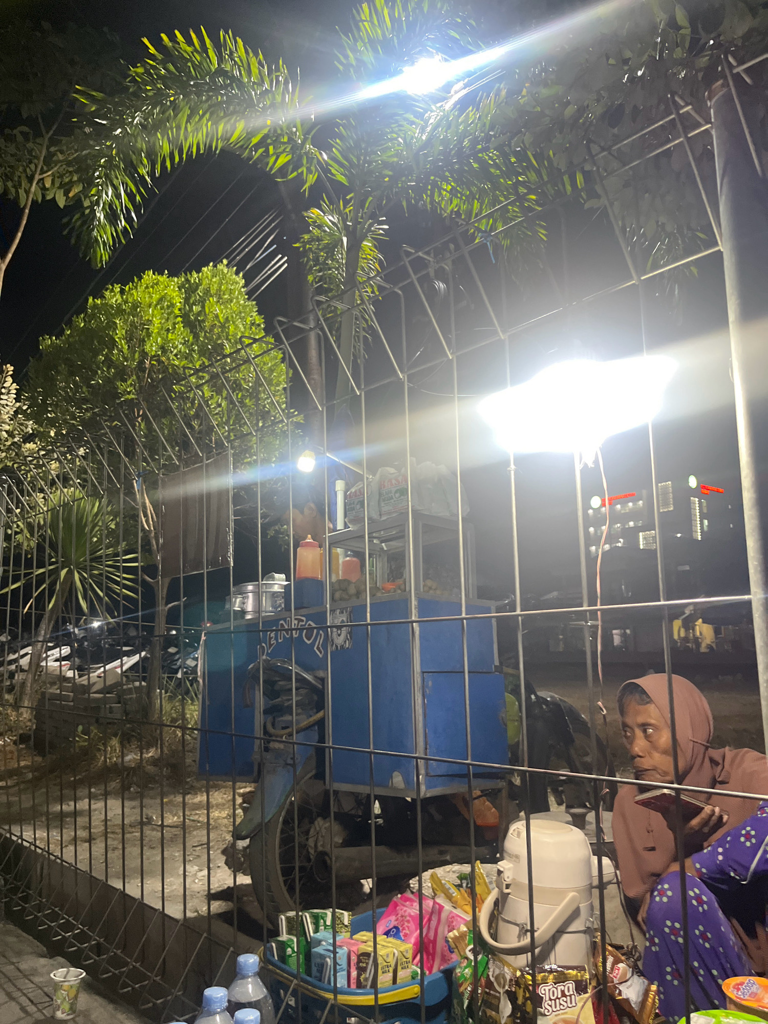 The image depicts a nighttime scene on the street in Banaran, Indonesia, likely a roadside stall or a vendor's cart. A woman, appearing to be the vendor, is seated on the right side of the frame, wearing a brown headscarf and a brown top, with purple patterned pants. She is holding a dark object, possibly a phone, in her hands and looking to her right. In front of her is a blue cart that has a motor vehicle attached, suggesting it's a mobile food or drink cart. The cart is stocked with various items, including bottles of drinks, packages of snacks, and a white container, possibly a thermos or dispenser. A bright light illuminates the scene from above, casting a glow on the surrounding foliage and the cart. Behind the cart, several parked motorcycles are visible, indicating a public area. In the background, a building with lit windows can be seen. The entire scene is enclosed by a metal fence, suggesting the stall might be within a designated area or has a protective barrier. The overall atmosphere is that of a typical night market or roadside vending area.
