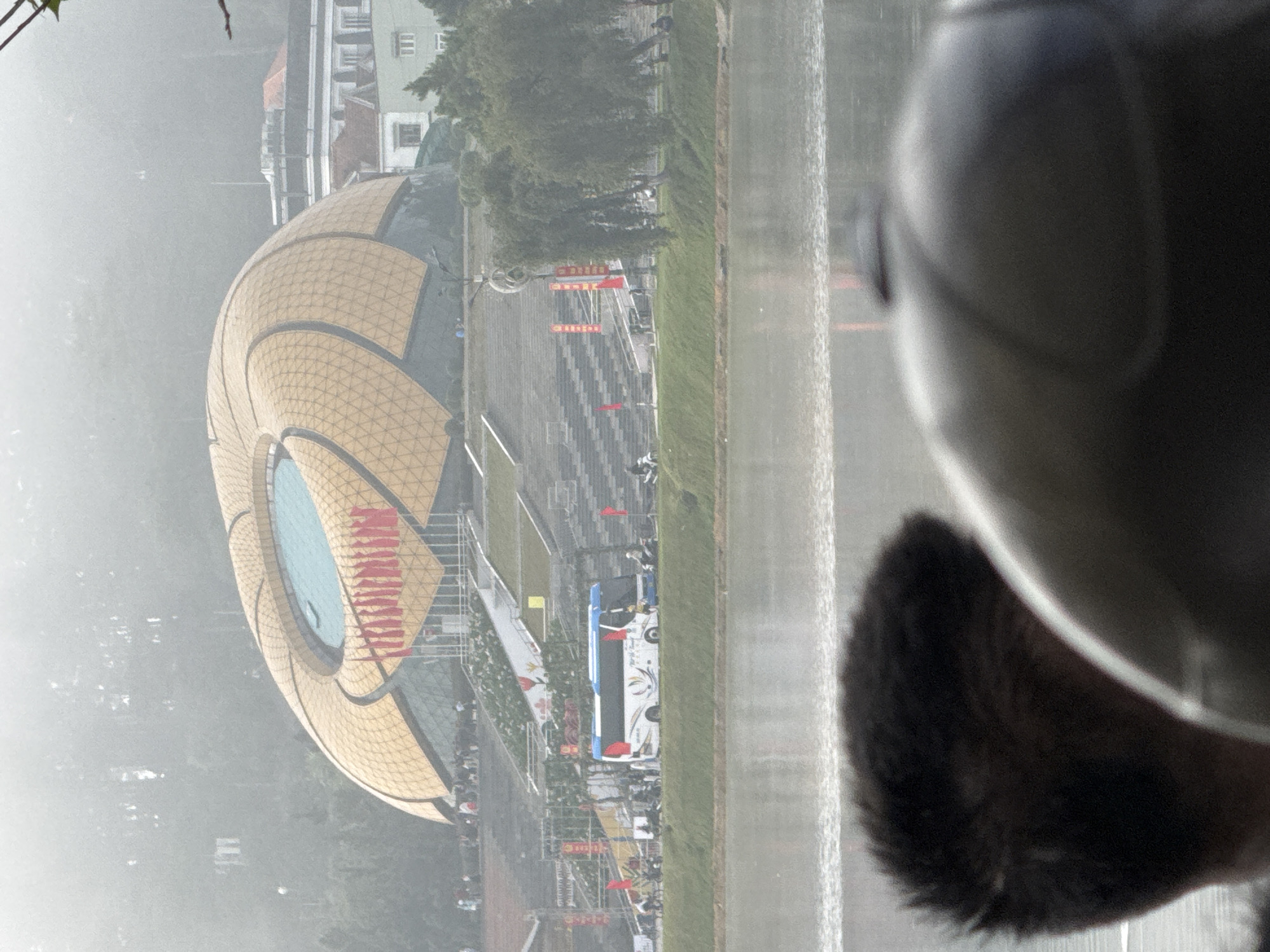 The image captures a misty view of the Da Lat Flower Garden, a prominent tourist attraction in Da Lat, Vietnam. In the foreground, blurred out, is the back of a person's head, potentially wearing a helmet. Beyond them, a body of water reflects the hazy sky. In the mid-ground, a large, unique geodesic dome structure, characteristic of the garden's architecture, stands prominently. Red flags are visible along the tiered steps leading up to the dome. A white bus with blue accents is parked on a road adjacent to the garden, and several motorbikes are visible. The background shows a lush, tree-covered hillside, shrouded in mist, with some buildings peeking through. The overall atmosphere is serene and somewhat obscured by the weather conditions.
