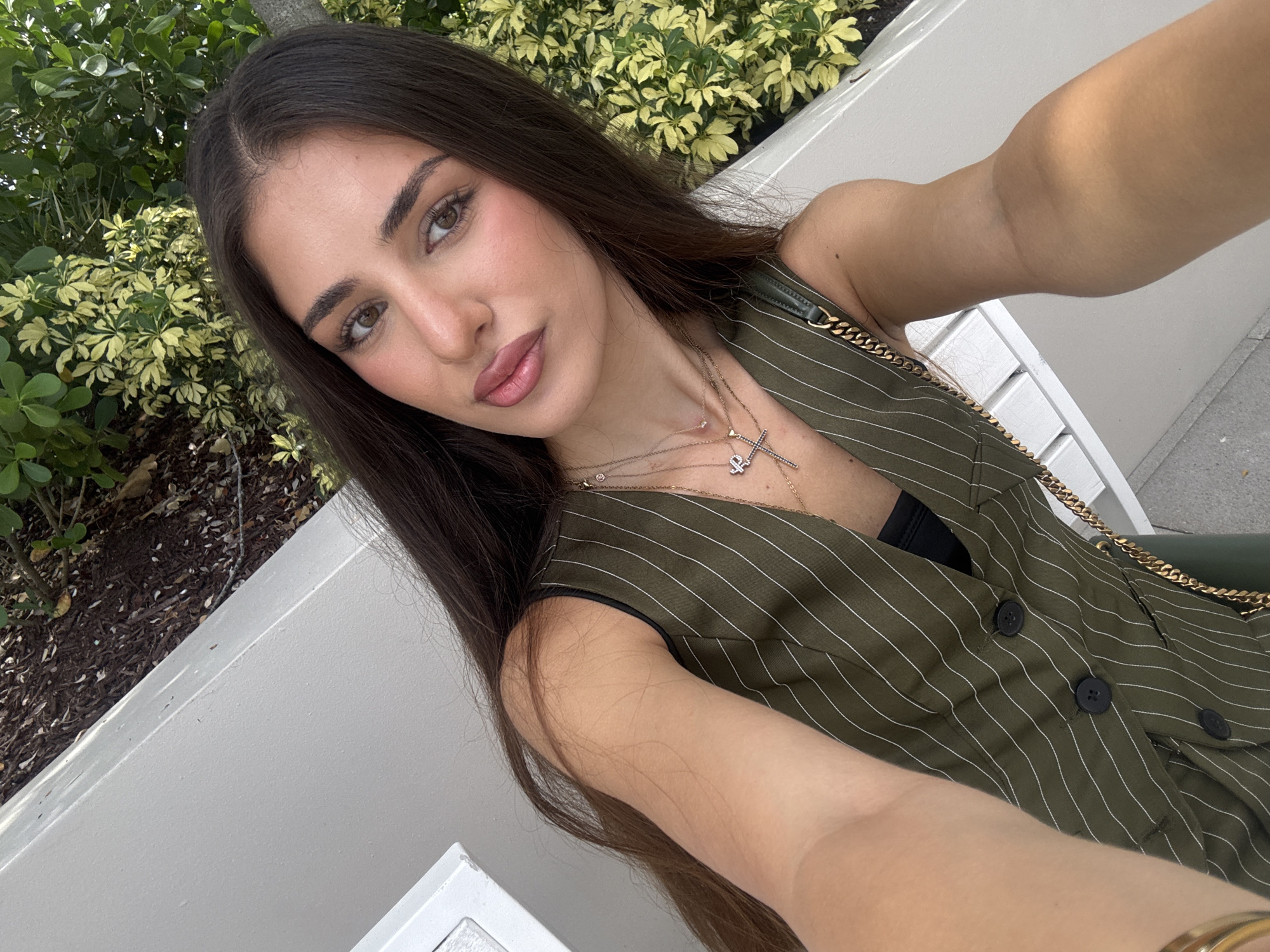 A young woman with long dark hair is taking a selfie outdoors. She is wearing a green pinstripe vest over a black top and several necklaces. She has light skin and is wearing makeup, including lipstick. Her arm is extended, holding the camera. Behind her, there is a light-colored bench, foliage with light green and yellow leaves, and dry ground cover. The setting appears to be a public outdoor space in Hallandale Beach, Florida, United States of America, possibly a park or garden area.