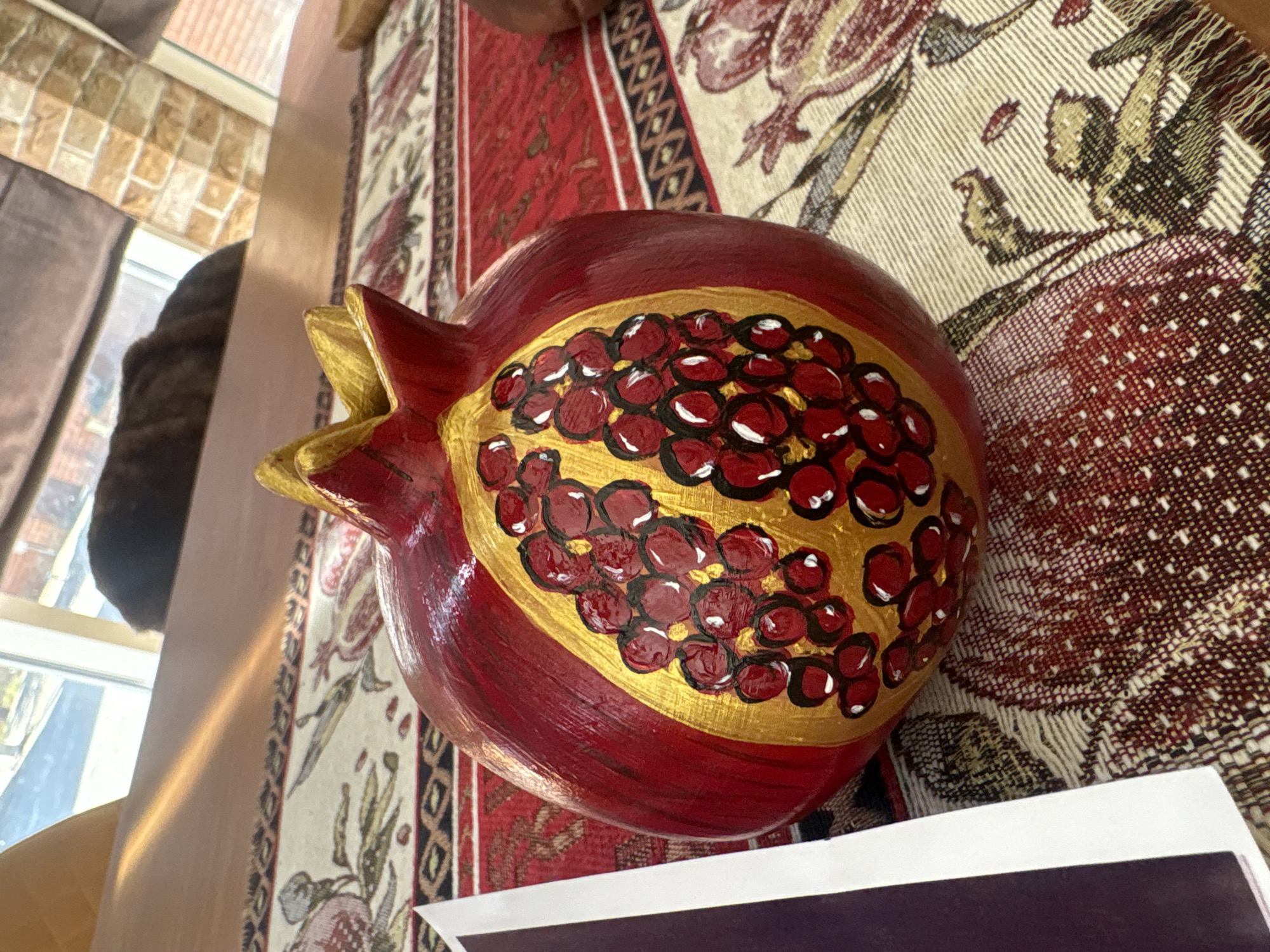An overhead, slightly angled, close-up shot features a decorative pomegranate, painted in vibrant red and gold, resting on a patterned tablecloth. The pomegranate is the primary subject, with its textured surface and painted arils clearly visible. It sits on a tablecloth with a complex pattern of red, cream, and dark brown, featuring floral and geometric motifs. To the left of the pomegranate, a portion of a white and dark purple card or paper is visible, partially overlapping the tablecloth. The background shows a wooden surface, likely a table, illuminated by natural light casting a diagonal glare. Further back, a window is visible, revealing a blurry outdoor scene with what appears to be a dark-colored animal, possibly a cat, lying down. The overall impression is one of indoor still life with a focus on the decorative pomegranate. This scene is not specifically located in Georgiyevsk, Russia, and there is no information provided to support such a claim.