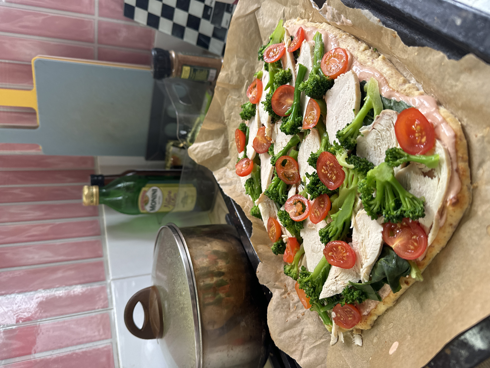 A partially eaten pizza sits on a parchment-lined baking sheet, featuring sliced chicken, broccoli florets, and halved cherry tomatoes. It appears to be located in a kitchen next to a cooking pot with a lid. A bottle of Monini olive oil is also visible near a cutting board and a spice container. The walls have pink and white tiles, topped with black and white checkerboard tiles. There is no indication that this picture was taken in Krasnoyarsk, Russia.