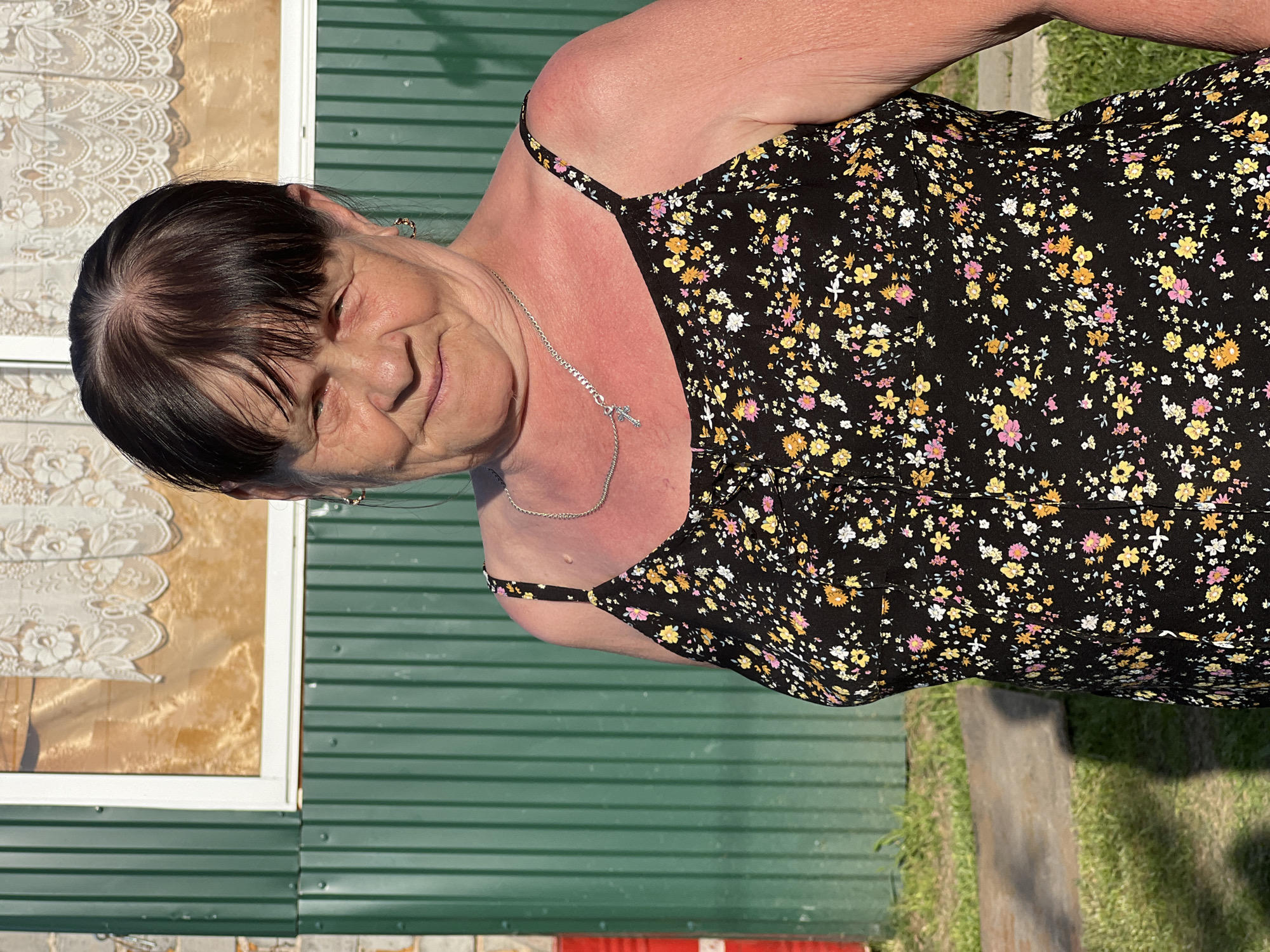 A woman is outdoors wearing a black floral sundress and a silver necklace with a cross pendant. She has short, dark hair with bangs, and appears to be middle-aged or older. The background features a green corrugated metal wall with a white-framed window that has lace curtains visible inside. The area seems to be a yard or garden, with some grass and a concrete walkway visible at the bottom of the frame. The location is in the city of Chaikovsky, Russia.