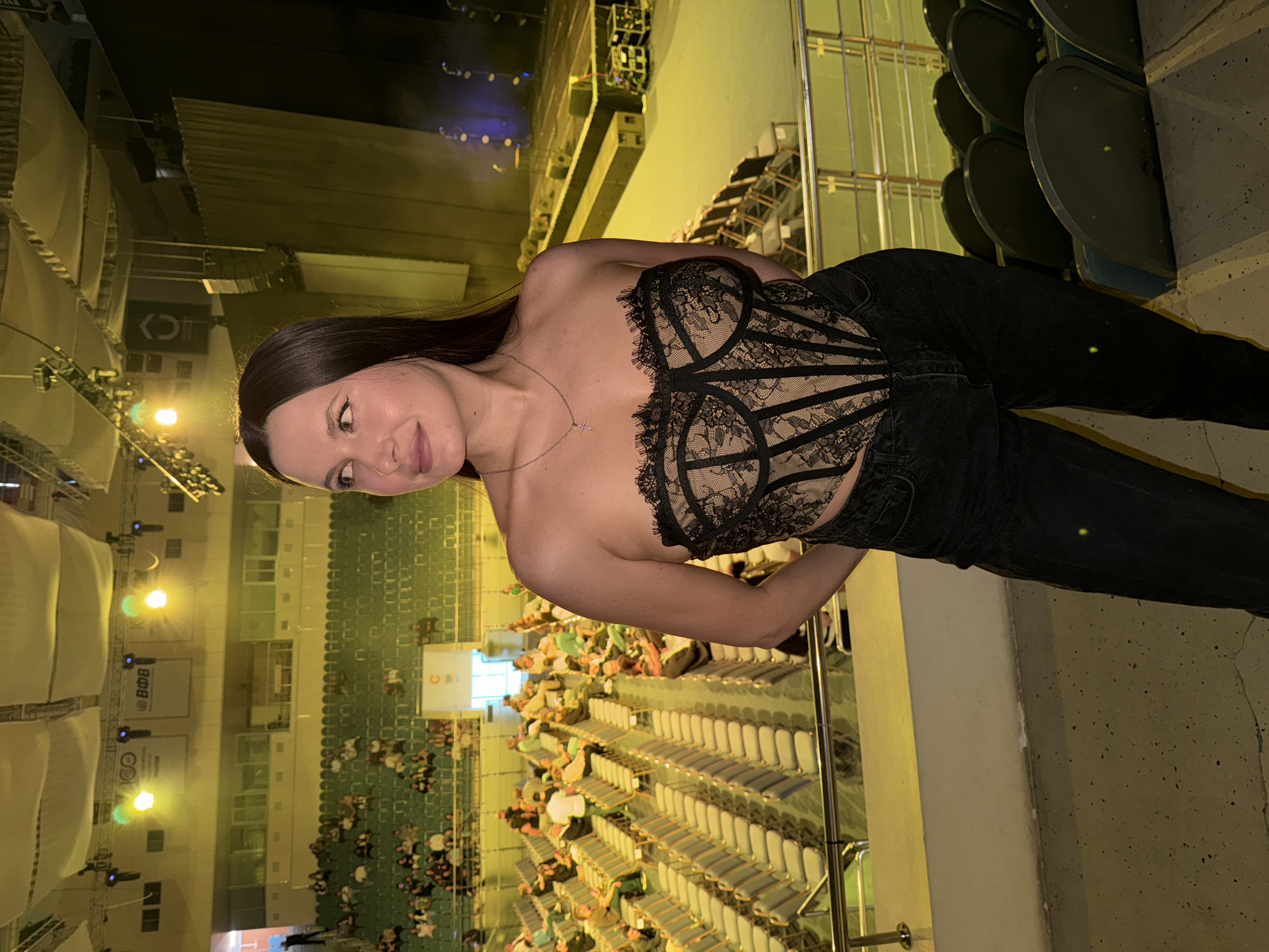 A waist-up shot captures a young woman posing inside a large, dimly lit indoor arena or auditorium. The image is presented sideways, with the woman on the left. She has long, straight dark hair, a light complexion, and a gentle smile, looking directly towards the camera. She is dressed in a sleeveless black lace corset-style top and black jeans, complemented by a delicate silver necklace with a small cross pendant.
The background reveals the tiered seating of the venue. Immediately behind her are several rows of empty dark-colored seats. Further back, numerous people are seated in the stands, suggesting an event is either about to commence or is already in progress. In the distance, a stage is visible, equipped with various lighting fixtures and structures, some casting spotlights. The ambient lighting is warm and yellowish, indicating an evening setting.
Several signs and banners are partially visible in the background. One white banner clearly displays the Cyrillic letters "ВФВ" (VFV). Another sign shows the number "100," while a stylized logo resembling "C>" and another text "co" or "oC" can also be seen. The scene suggests a public event such as a concert, performance, or a large gathering. The woman appears calm and relaxed.