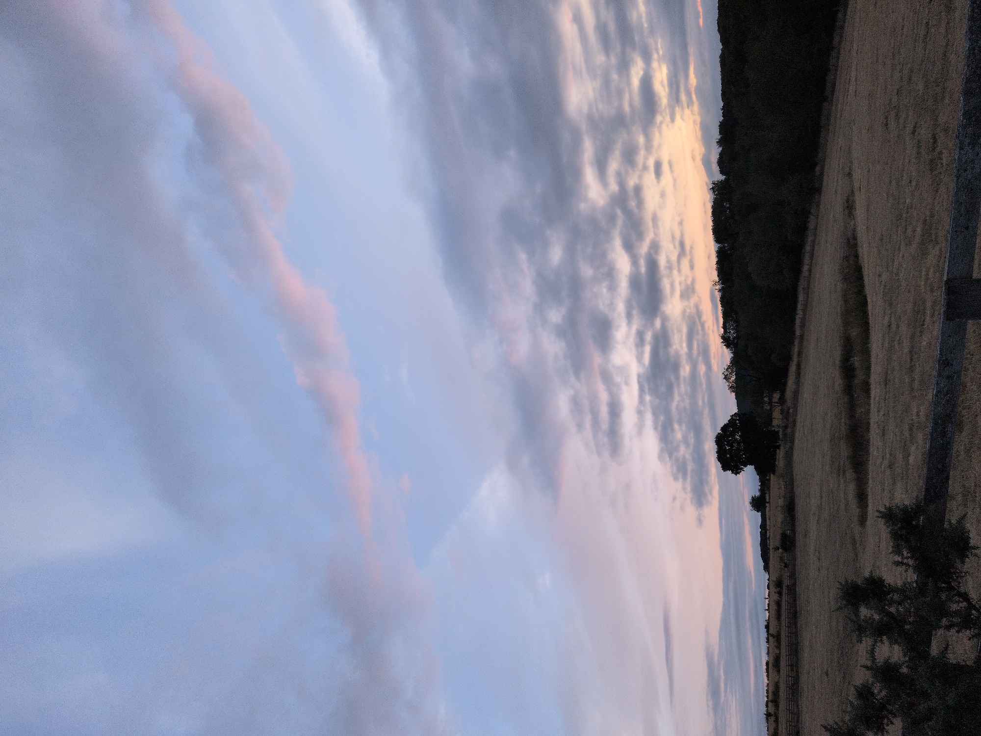 The image captures a rural landscape in Little Wenlock, United Kingdom, under a twilight sky. The sky is a blend of soft blues and pale pinks, with wispy clouds streaking across. Below, a field of dry, golden grass stretches out, marked by a low wooden fence in the foreground. A solitary, dark tree stands prominently in the middle ground, silhouetted against the fading light. Behind the field, a dense line of dark trees forms the horizon, suggesting a wooded area. The overall impression is one of peaceful natural scenery at the close of day.