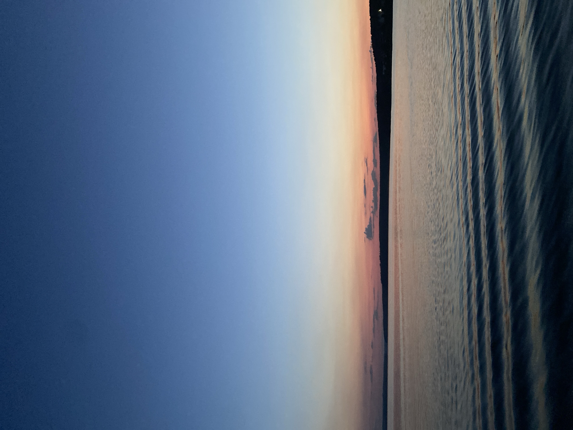 This is a photograph of a tranquil body of water with a silhouetted tree line in the distance, set against a twilight sky. The sky transitions from a deep blue at the top to softer hues of pink and orange near the horizon, indicating the time of day is likely just after sunset or before sunrise. The water exhibits gentle ripples, reflecting the colors of the sky. The far shore is a dark, indistinct line of trees and possibly some structures, hinting at a natural landscape. The image captures a serene and calm atmosphere. This scene is likely depicting the Kama River in the city of Votkinsk, Russia, given the typical geography and natural scenery found in that region during twilight. There are no people or specific actions depicted in the image.