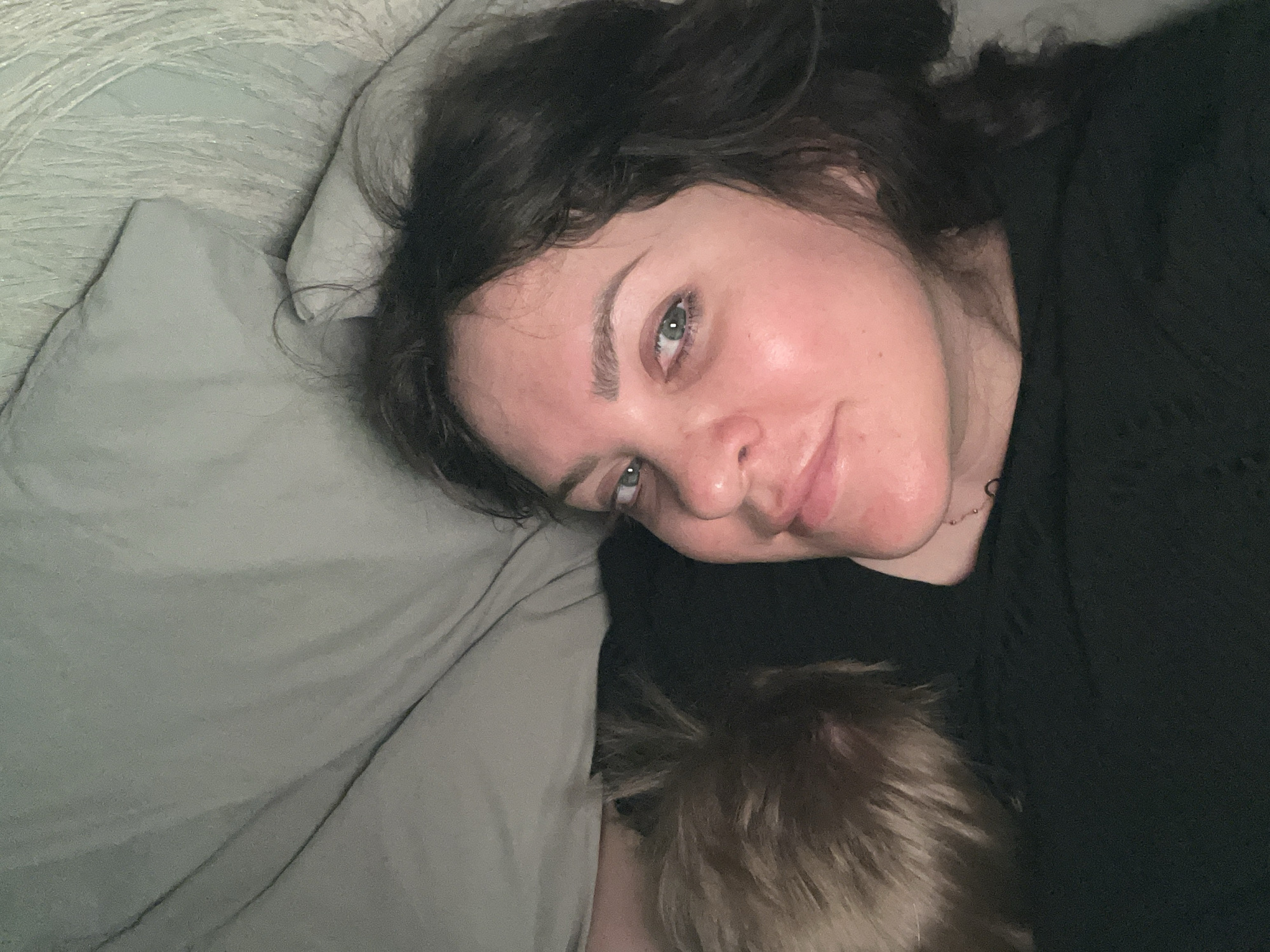 A person with dark hair and green eyes is lying on a gray pillow and blanket. Part of a child's head with blonde hair is visible near the person's chest. The person is wearing a black, textured shirt. The background appears to be a bed in a room. There is no evidence of the city of Noviy, Russia.