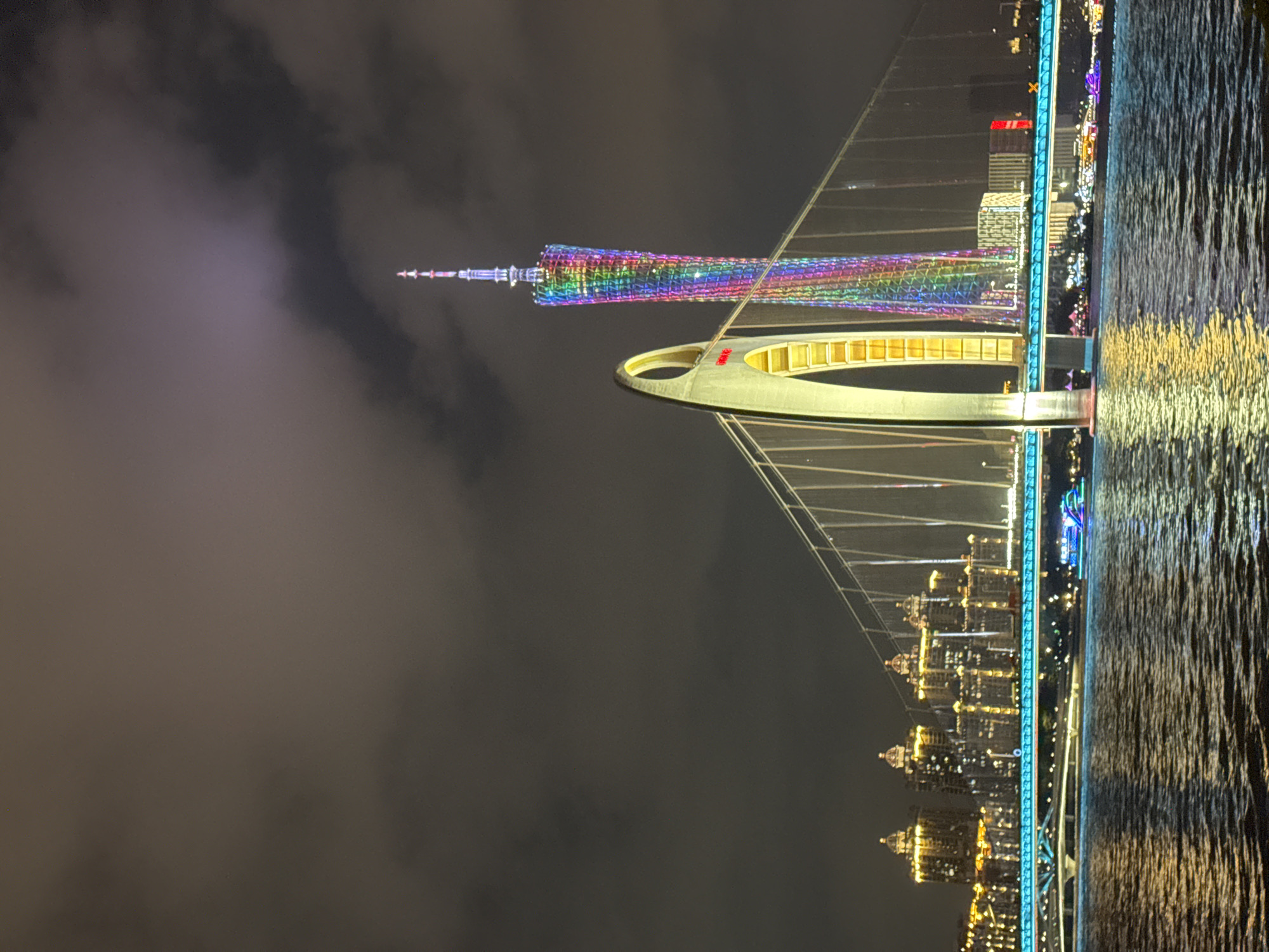 The image shows a nighttime view of the Canton Tower and a nearby bridge in the Tianhe District of Guangzhou, China. The Canton Tower is illuminated with colorful lights. A bridge with a distinct, curved design spans a body of water, reflecting lights from the city and the bridge itself. Other buildings and city lights are visible in the background, along the waterfront. The sky is dark and cloudy.