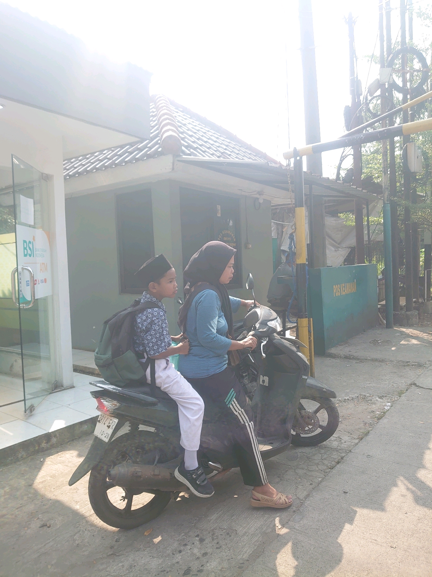 In Bojong Gede, Indonesia, two individuals are seen on a motorcycle in front of a building identified as "POS KEAMANAN". A young boy, wearing a black cap, a patterned shirt, white pants, and a backpack, is seated behind a woman. The woman is wearing a black hijab and a blue striped shirt with black athletic pants. She is operating the motorcycle, which is dark green and has a license plate that reads "B 3114 EIX 10-28". The background features a building with a tiled roof, and part of what appears to be an ATM area for BSI Bank is visible to the left. The scene is outdoors, with bright sunlight casting shadows on the ground, suggesting it is daytime. The overall environment appears to be a public or semi-public space within a town.