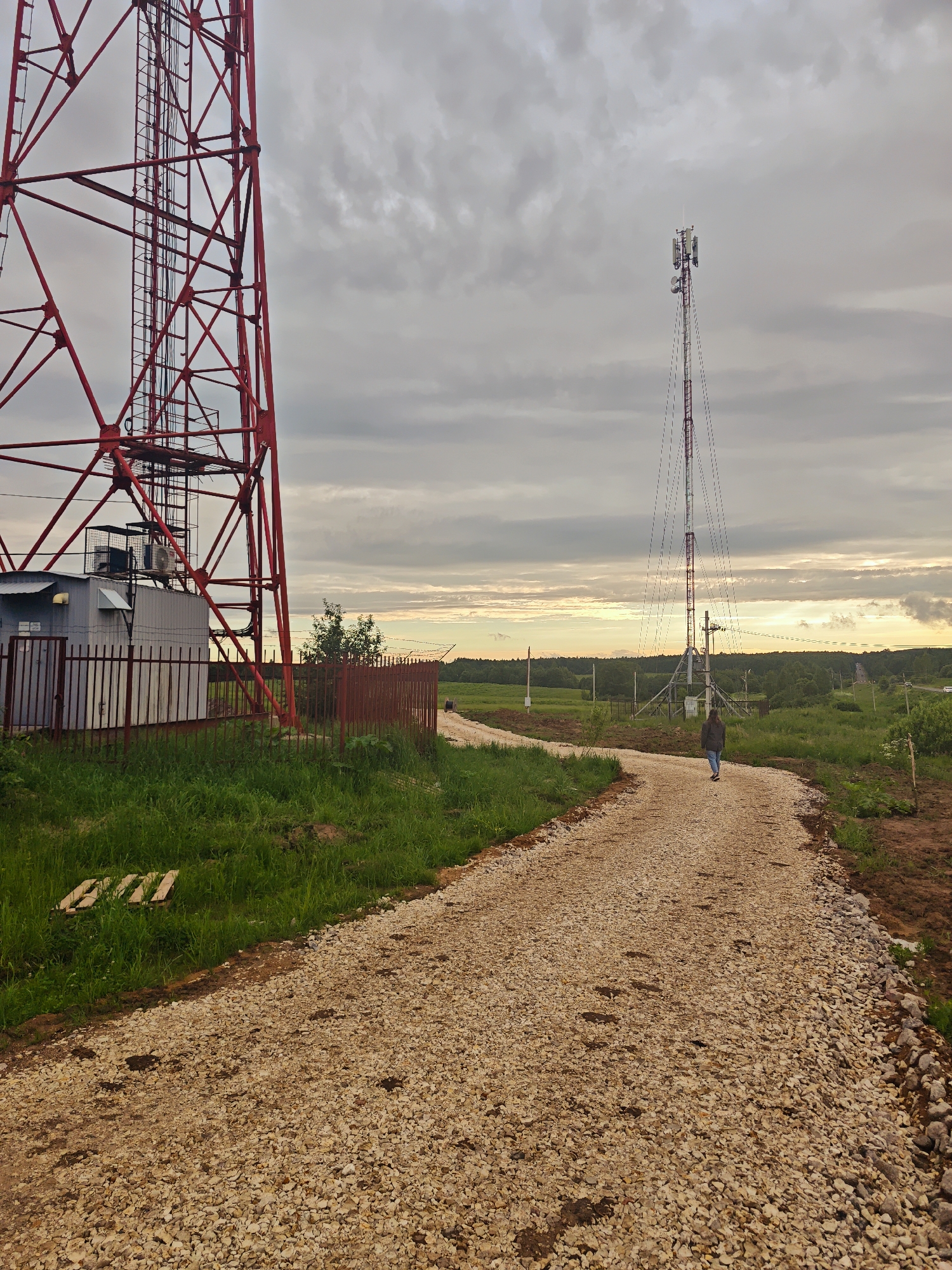 An overcast sky looms over a rural landscape in Fedorovskoe, Russia, featuring two communication towers and a lone person walking down a gravel path. The foreground is dominated by a large, red lattice tower on the left, with a small, grey building enclosed by a fence at its base. A winding gravel path stretches into the middle ground, leading towards a second, taller communication tower. A person, viewed from behind, walks along this path. The background consists of green fields, scattered trees, and more distant trees forming a horizon line. The scene is captured from a slightly low angle, emphasizing the scale of the towers.