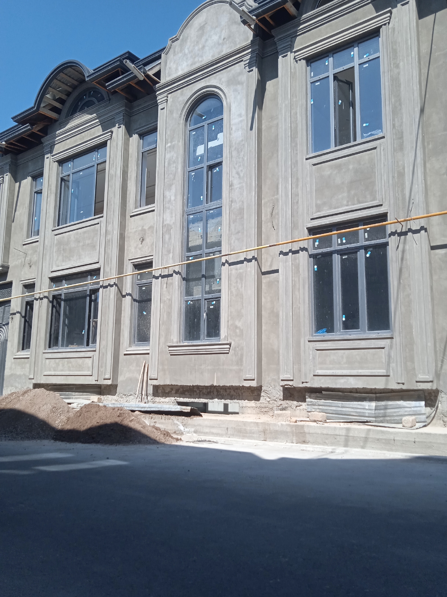 The image shows the exterior of a grey building in Eshanguzar, Uzbekistan. The building appears to be under construction or recently plastered, with unfinished ground surrounding its base. The building has several large windows, some rectangular and one arched, with grey frames. A yellow gas pipe runs horizontally along the facade. A pile of dirt is located in front of the building, and stacks of grey sheets are stacked up against the building. The street is visible in the foreground, partly in shadow. The sky is clear blue.