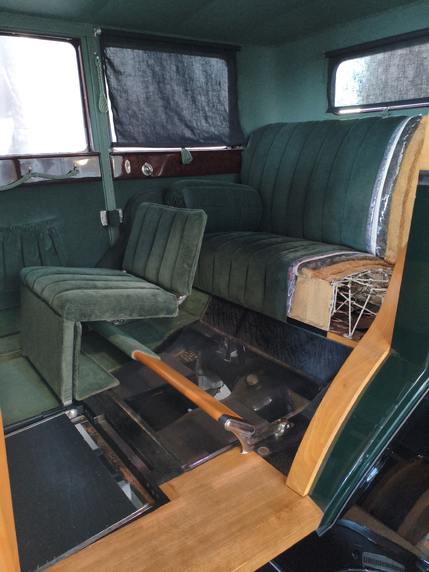 The image shows the interior of a vintage car, possibly a 1920s or 1930s model, with dark green velvet upholstery. The scene is indoors, with natural light coming from the left side, illuminating the interior.
In the foreground, a wooden plank runs diagonally across the bottom of the image. To its left, a black surface with metal trim suggests a floor mat or panel. Further back, a smaller, dark green velvet seat is visible, with a wooden lever extending from its front and angled downwards.
The main seating area features a bench seat upholstered in dark green velvet with a distinct channel tufting pattern. The side of the seat is partially cut away, revealing its internal structure, including springs, burlap, and padding. This cutaway reveals the construction of the seat, indicating it is on display or undergoing restoration.
Behind the bench seat, a dark fabric curtain is drawn across a window. Another window, to the left, is partially covered by a similar curtain, with bright light filtering through. The car's interior walls and ceiling are also covered in dark green fabric. The overall impression is of a well-preserved, possibly museum-quality vehicle.
The context of Ingolstadt, Germany, is not directly evident from the visual information within the image itself. However, assuming this car is located in Ingolstadt, it is likely part of a museum collection, a private collection, or a restoration facility within the city.