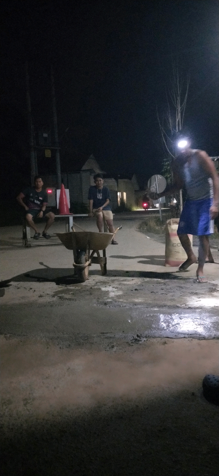 In Belimbing Raya, Indonesia, at night, four individuals are present at what appears to be a construction or road repair site. Two men are seated on a bench, one on the left wearing a dark t-shirt and shorts, and another on the right wearing a dark t-shirt and light-colored shorts. A third man, wearing a grey tank top and blue shorts, is standing to the right and holding a circular object, possibly a sign, and has a headlamp illuminating his face. A fourth individual is also visible in the distance on the far right, partially obscured. In the foreground, a wheelbarrow is positioned on a wet, uneven surface that looks like wet cement or mud. A large bag, possibly containing cement, is situated behind the standing man. A red traffic cone is placed on a metal structure behind the seated men. The environment suggests an outdoor, possibly rural or suburban, setting with minimal ambient light, with the primary illumination coming from artificial sources like streetlights and the headlamp.