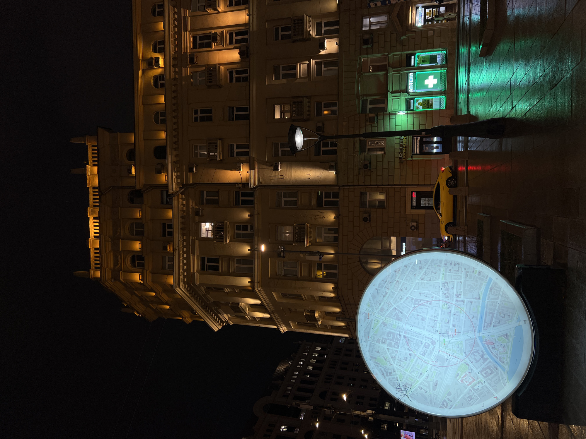 At night, the city of Moscow, Russia, features a large, illuminated circular map on the sidewalk. A yellow taxi is parked nearby. A building with lighted windows rises in the background, with a green pharmacy sign visible on one of its storefronts. A lamppost extends out over the sidewalk.