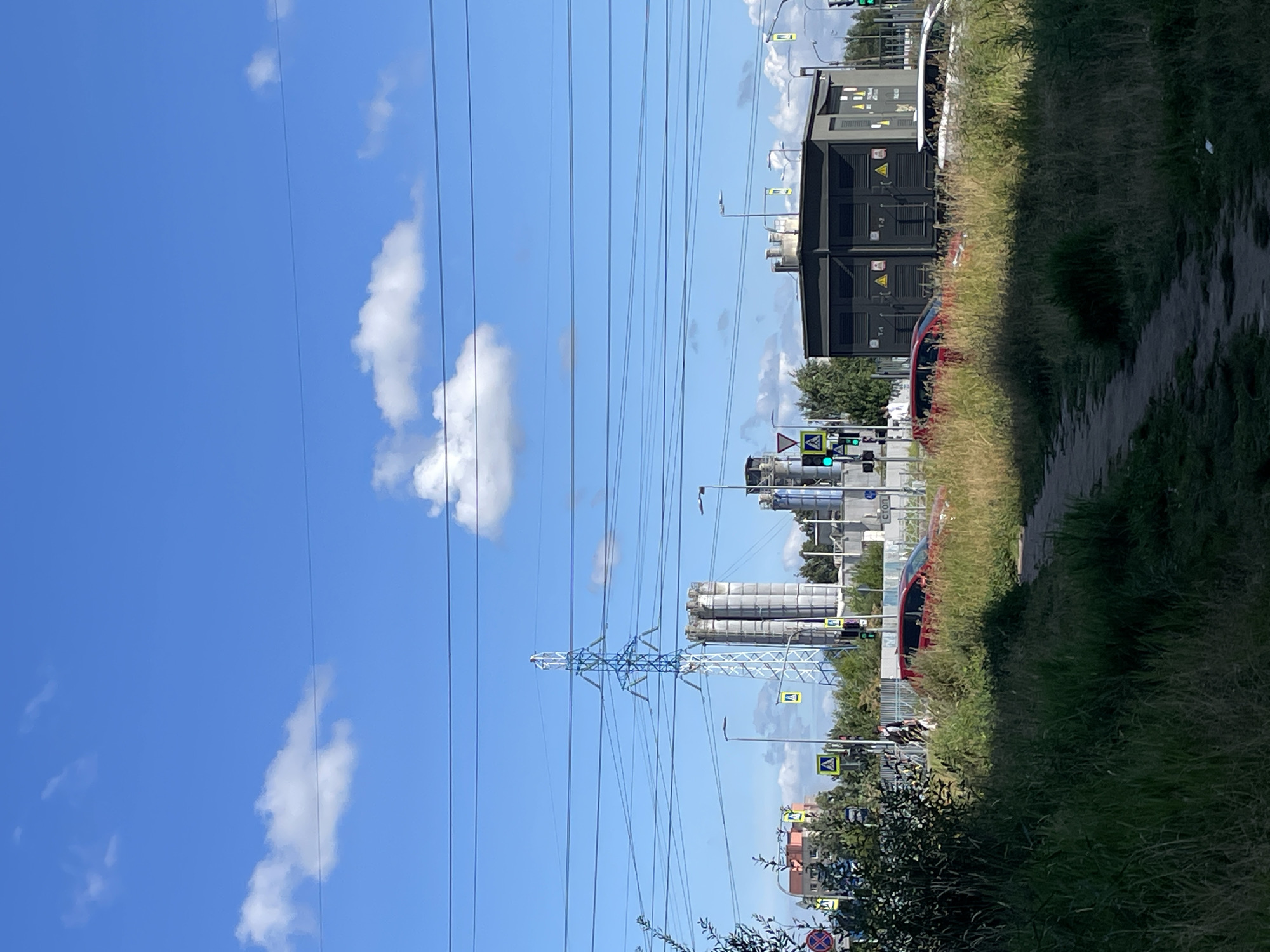 In an urban landscape under a bright blue sky with scattered white clouds, a scene unfolds featuring industrial structures and passing vehicles. Dominating the background are several tall, cylindrical concrete silos, with a prominent lattice metal power pylon positioned to their left, its white and blue frame supporting a network of overhead power lines that stretch across the frame. To the right of the silos, a dark, rectangular industrial building with visible vents stands adjacent to a road.
In the mid-ground, a red car is visible driving on the road, while further down the road, a white vehicle is also in motion. The road is lined with traffic lights, some displaying green signals, and pedestrian crossing signs featuring a yellow triangle with a black pedestrian symbol. A person is seen walking on the sidewalk near the red car.
The foreground is characterized by a path running through tall, dry grass and vegetation, suggesting a somewhat unkempt or natural area within the city. Trees with green foliage are scattered throughout the scene, particularly behind the industrial buildings. The overall impression is one of a functional, possibly industrial or infrastructure-heavy area in a city on a clear day.