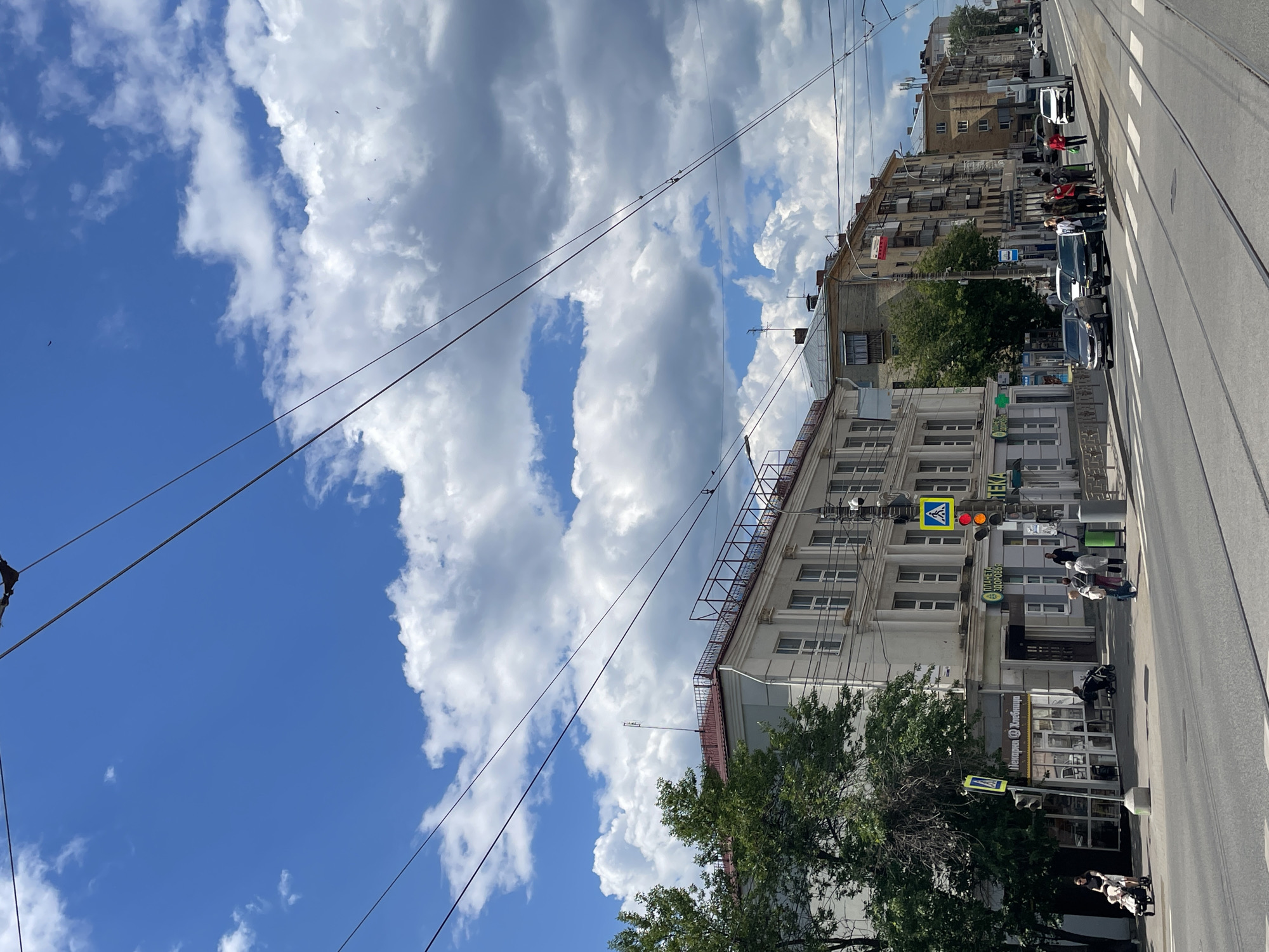 A street scene unfolds in a city under a partly cloudy sky. Overhead, power lines crisscross the frame, suggesting an urban environment. On the left, a light-colored, multi-story building with white columns and numerous windows lines the street. A large tree with green foliage stands in front of it. A pedestrian crossing is marked on the road. A woman pushes a stroller on the sidewalk, and another person walks with a stroller. Traffic lights are visible, with one showing red.
Further down the street, more buildings, appearing to be residential with balconies, are visible. People are gathered at a bus stop, waiting for transportation. Several cars are on the road, and a white truck is moving away from the camera. The street has tram tracks running through it. The overall impression is of a typical urban street with everyday activities taking place.