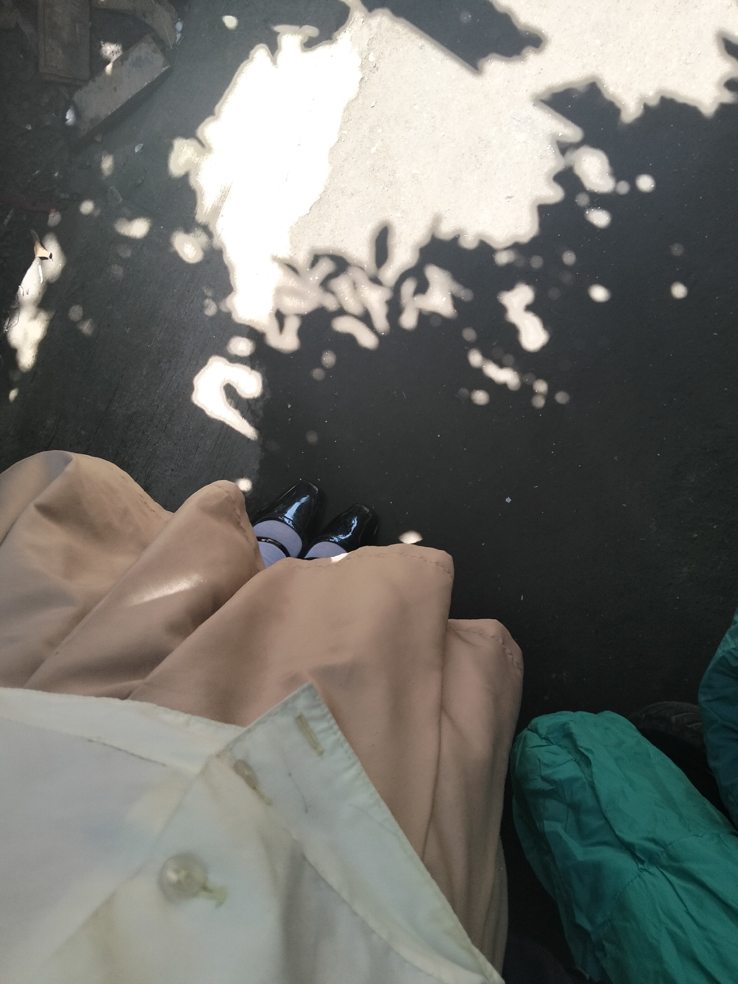 In an outdoor urban setting, possibly a sidewalk or paved area in Davao City, Philippines, the image captures a close-up perspective of a person's lower legs and feet. The ground is dark asphalt, partially illuminated by dappled sunlight casting shadows from overhead foliage or structures, creating bright, irregular patches and smaller circular spots of light.
The individual is wearing dark, glossy shoes, likely dress shoes or heels, with a portion of their feet visible. They are also clad in clothing that includes a light beige, possibly fabric, garment that drapes prominently in the foreground. Beneath this, a white collared shirt or blouse is visible, with a button and stitching details. To the right of the beige garment, a bright green, crinkled fabric object, perhaps a bag or another piece of clothing, is partially visible.
The overall impression is of a person standing or walking in an area with contrasting light and shadow. The specific location within Davao City is not definitively indicated by any landmarks or recognizable features in the image.
