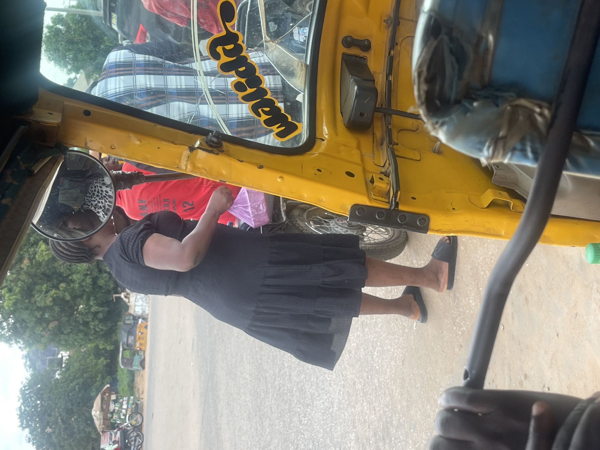 A woman in a black dress and black sandals stands next to a yellow auto-rickshaw in what appears to be an outdoor market area in Abuja, Nigeria. The auto-rickshaw has a sticker on its windshield that reads "Walida." In the background, there are other vehicles, including a motorcycle, and market stalls with various goods. The woman is holding a pink plastic bag. The scene is captured from inside another auto-rickshaw, suggesting the photographer is a passenger. The lighting indicates it is daytime.