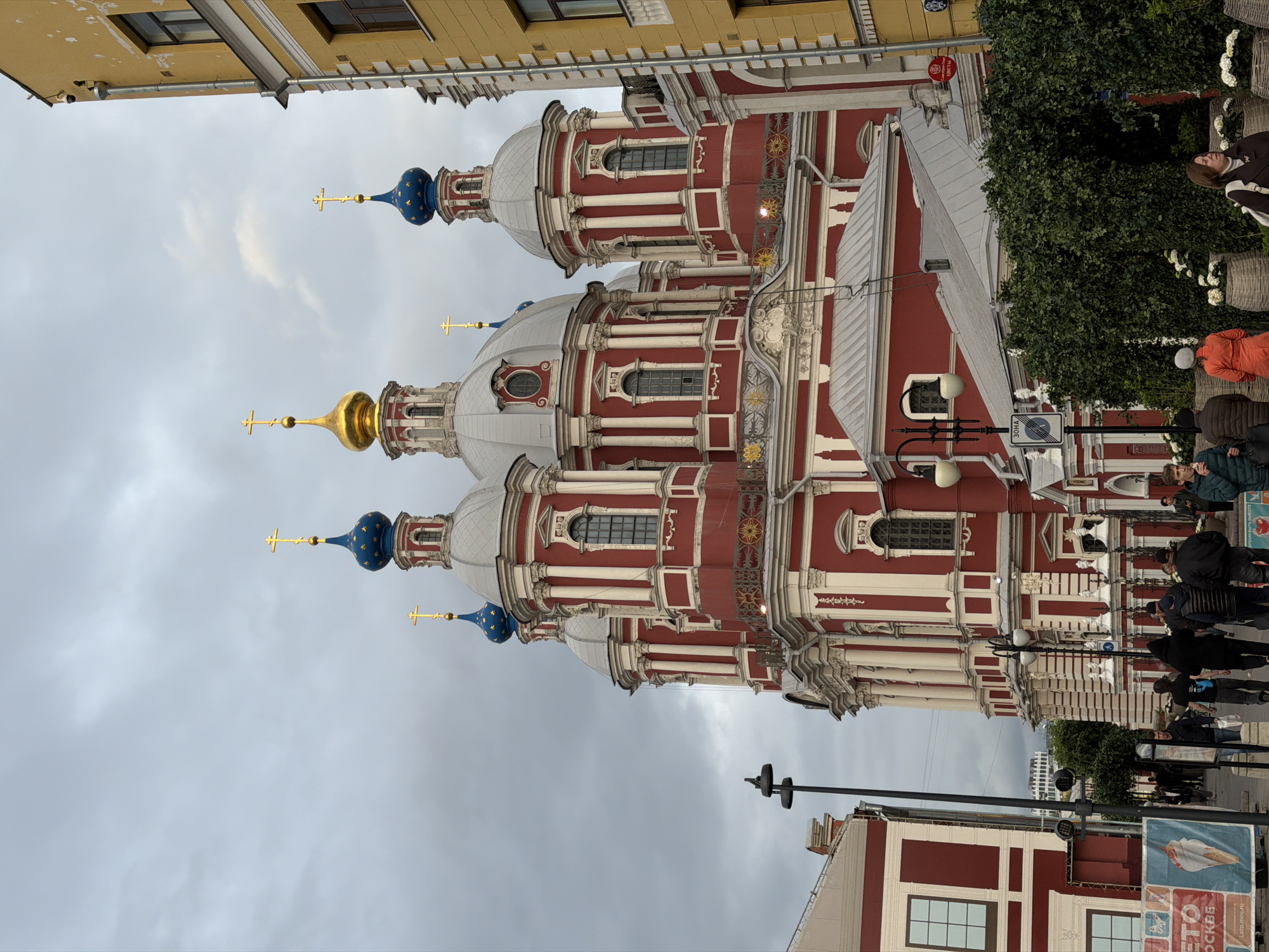 The image depicts the exterior of the Church of the Life-Giving Trinity in Moscow, Russia. The church has a red and white facade with multiple domes, one of which is gold and the others are blue with gold stars. People are walking along the sidewalk in front of the church. Buildings and trees are visible in the background. The sky is overcast. Streetlights and signs are also visible in the foreground.