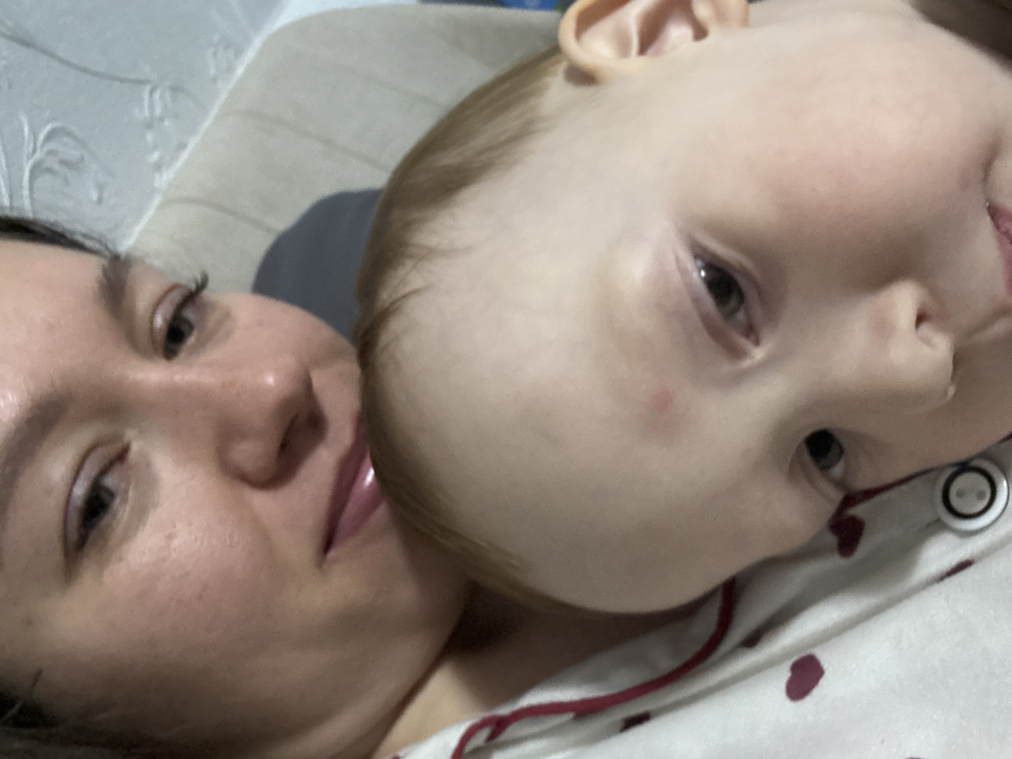 A close-up, tilted shot shows a woman and a young child lying together. The woman's face, positioned to the left, is partially visible, focusing on her eye, nose, and lips. The child's head is positioned on the right, with their ear visible. The subjects are lying on a patterned fabric with red heart shapes. The background is blurred, but a textured white surface is present. Location is uncertain based on the given image.