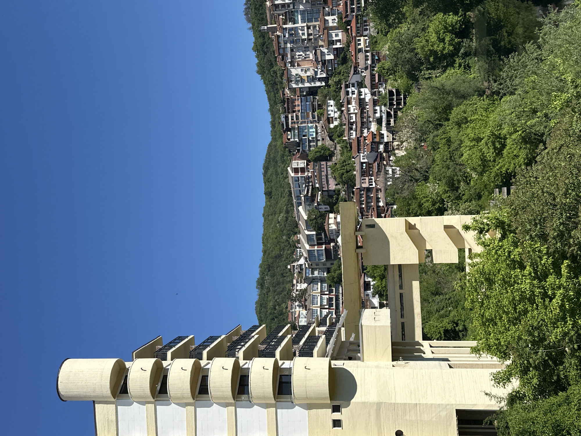 The image shows a building with a distinctive architectural style in Veliko Tarnovo, Bulgaria. The building has a facade with repeating cylindrical elements and open balconies, predominantly in white and beige tones. Behind the building is a hillside densely populated with houses and vegetation. The sky is clear blue. There are no people visible in the image.