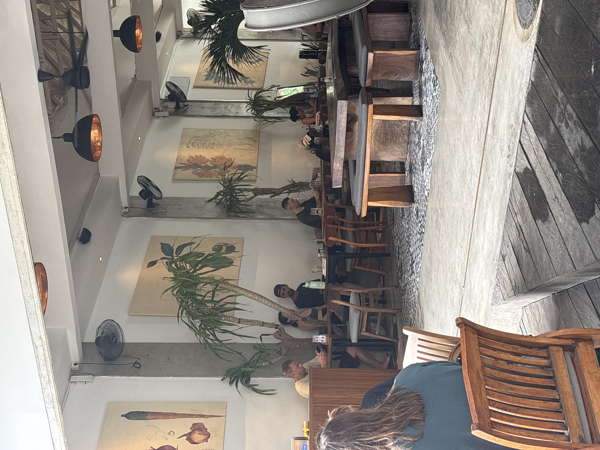 The image captures the interior of a spacious, open-plan restaurant or cafe, likely located in Kuta Selatan, Indonesia, given the context. The perspective is tilted, looking across a dining area with multiple patrons.
Several people are seated at wooden tables, engaged in dining, drinking, or using their mobile phones. In the foreground, the back of a person with long, curly brown hair is visible, seated at a wooden table with a matching chair. Further into the scene, various individuals are seated at long communal tables made of dark wood with light-colored cushions on the chairs. One man with a beard and a baseball cap is seen looking down, a water bottle next to him. Another man in a light-colored shirt is also visible, looking at his phone.
The establishment features a modern and somewhat industrial design, characterized by high white ceilings, exposed concrete beams, and white walls. Tropical palm-like plants in pots are strategically placed throughout the dining area, adding a natural element. The walls are adorned with framed botanical art prints depicting flowers and roots. For lighting and cooling, the space utilizes black, industrial-style pendant lights with a warm orange interior glow, as well as multiple black pedestal fans placed on ledges and ceiling fans. The flooring transitions between a tiled pattern and dark wooden planks.
The ambiance appears relaxed and casual, with people enjoying their meals and company. No specific text is clearly decipherable within the image.