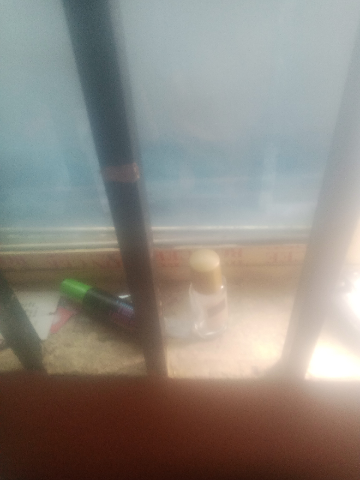 In a dimly lit indoor scene, a dark cylindrical object, possibly a pen or marker, with a gold band around its middle, stands vertically. To its left and slightly in front, a green-capped marker lies horizontally on a surface. Further to the right and slightly behind the green marker, a small clear glass bottle with a gold cap is visible. The background appears to be a window with a faint blue hue, suggesting it might be daytime. The overall scene is blurry, with some light glare obscuring finer details.