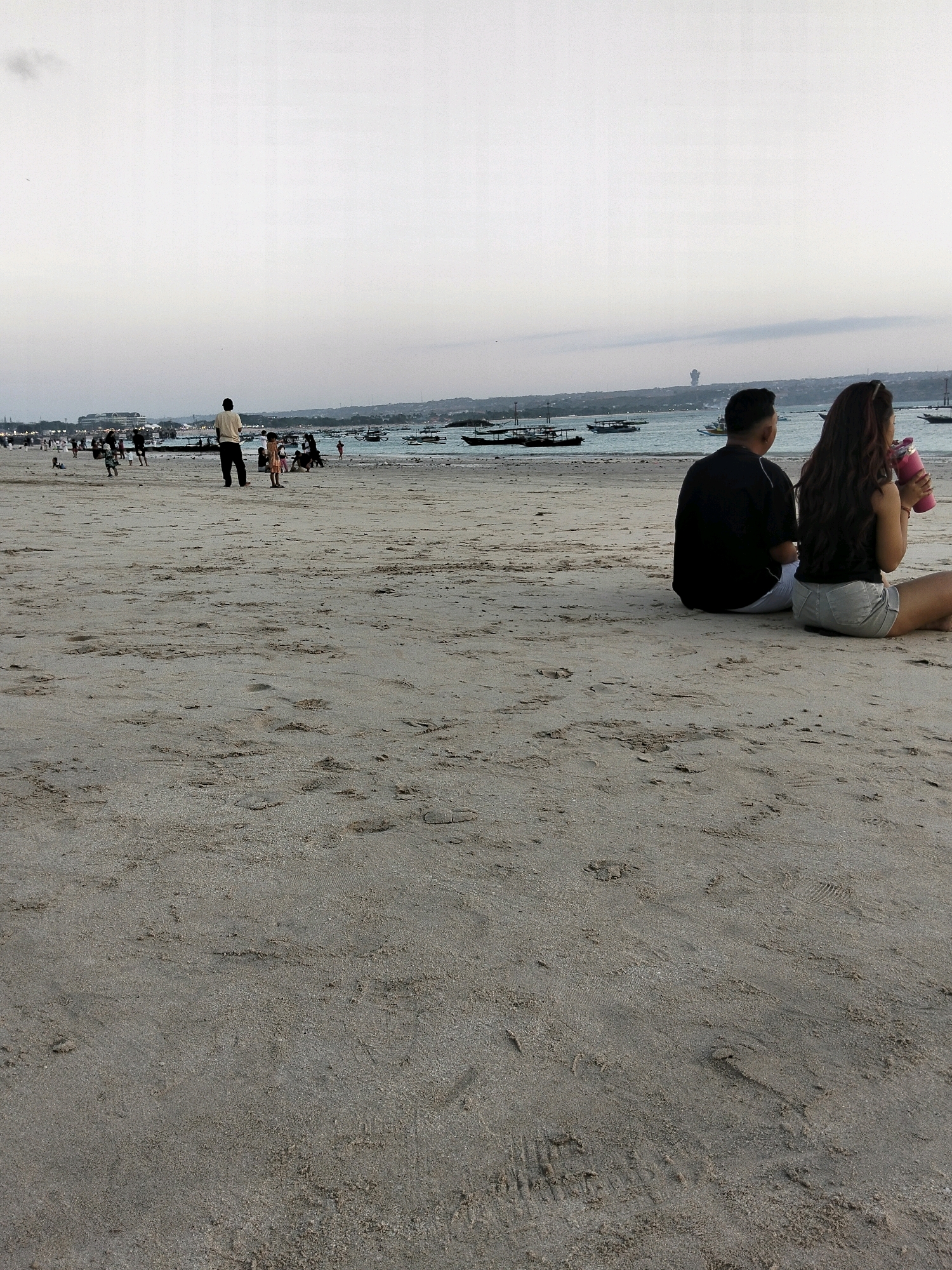 The image depicts a sandy beach in Denpasar, Indonesia, likely Kuta Beach due to the presence of numerous boats offshore and the general atmosphere. In the foreground, two individuals, a man and a woman, are seated on the sand, facing away from the camera and towards the ocean. The woman is holding a pink beverage container. In the middle ground, a group of people are visible on the beach, including a man in a light-colored shirt and dark pants standing and facing the water, and several other figures engaged in various activities. The background shows a calm body of water with many traditional Indonesian outrigger boats (prahu) anchored or sailing, and a distant coastline with a hazy sky. The overall lighting suggests it is late afternoon or early evening.