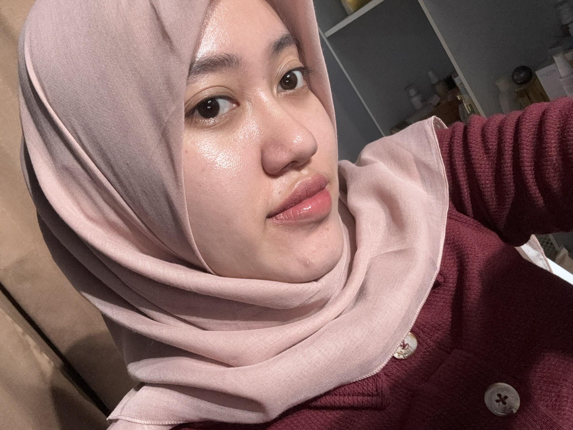 A close-up self-portrait of a person wearing a light pink hijab and a maroon cardigan. The person has fair skin, dark eyes, and glossy lips. The background is blurry, showing a white shelf with various containers. There is no indication of Gadingrejo, Indonesia.