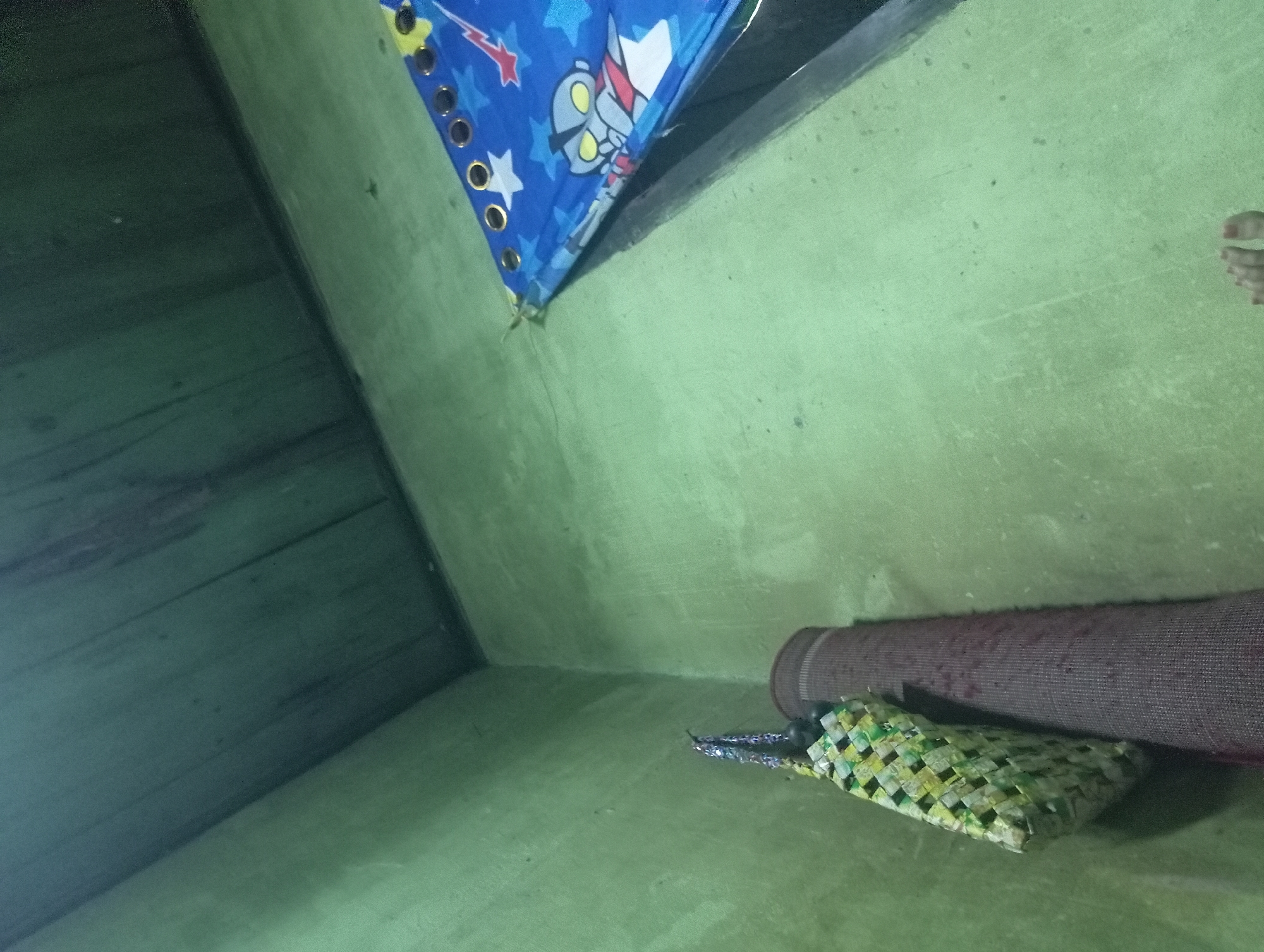 In a room with light green walls and a dark wooden ceiling, a woven bag with a checkered pattern of yellow and green hangs from a hook. To its right, a rolled-up rug with a dark maroon color stands upright against the wall. In the upper right corner, a blue fabric with white stars and a cartoon character is draped, showing grommets along its edge. The floor appears to be a light green surface, and in the bottom right of the frame, a person's bare feet are visible. The scene suggests an indoor residential setting, possibly a bedroom or storage area, in the city of Rejosari, Indonesia.