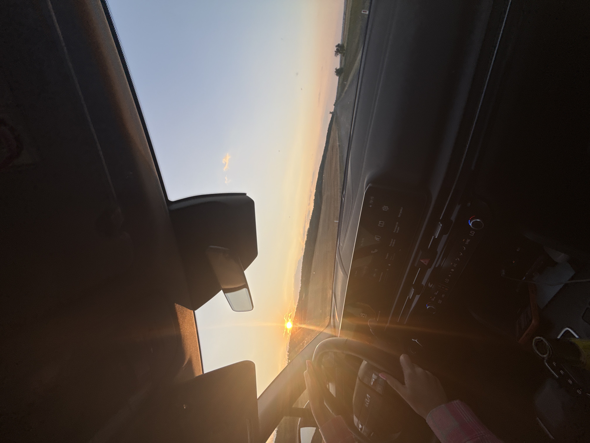 The image is taken from inside a car, looking out at a sunset. A person's hands are on the steering wheel. The dashboard of the car is visible, with a digital display showing the time as 19:15. The setting is rural, with fields and trees visible outside the car. The sun is low on the horizon, casting a warm glow. There is no explicit information to confirm the location as Sadaclia, Moldova, but the description fits a scene that could be found in such a location during sunset.