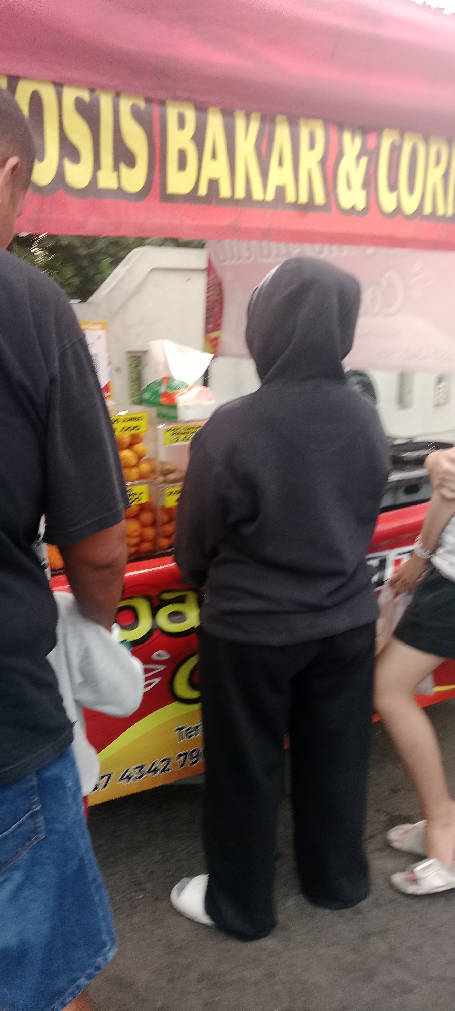 An outdoor scene in Ngemplak, Indonesia, depicts a food stall with a red and yellow awning displaying text in yellow with a black outline, partially visible as "OSIS BAKAR & CORN". Several people are present. On the left, a person wearing a black t-shirt and blue jeans is partially visible from the back, holding a white cloth. In the center, a person in a black hooded sweatshirt and black pants is standing. To the right, a person wearing a white and grey striped shirt and a black skirt is visible from the knees down, wearing white sandals. The stall itself has clear plastic containers holding orange fruits, likely oranges, and other items. The ground appears to be paved. A red banner on the stall has yellow lettering, including the partial text "ga" and "C". Further down the red banner, yellow lettering includes numbers that appear to be part of a phone number: "7 4342 79".