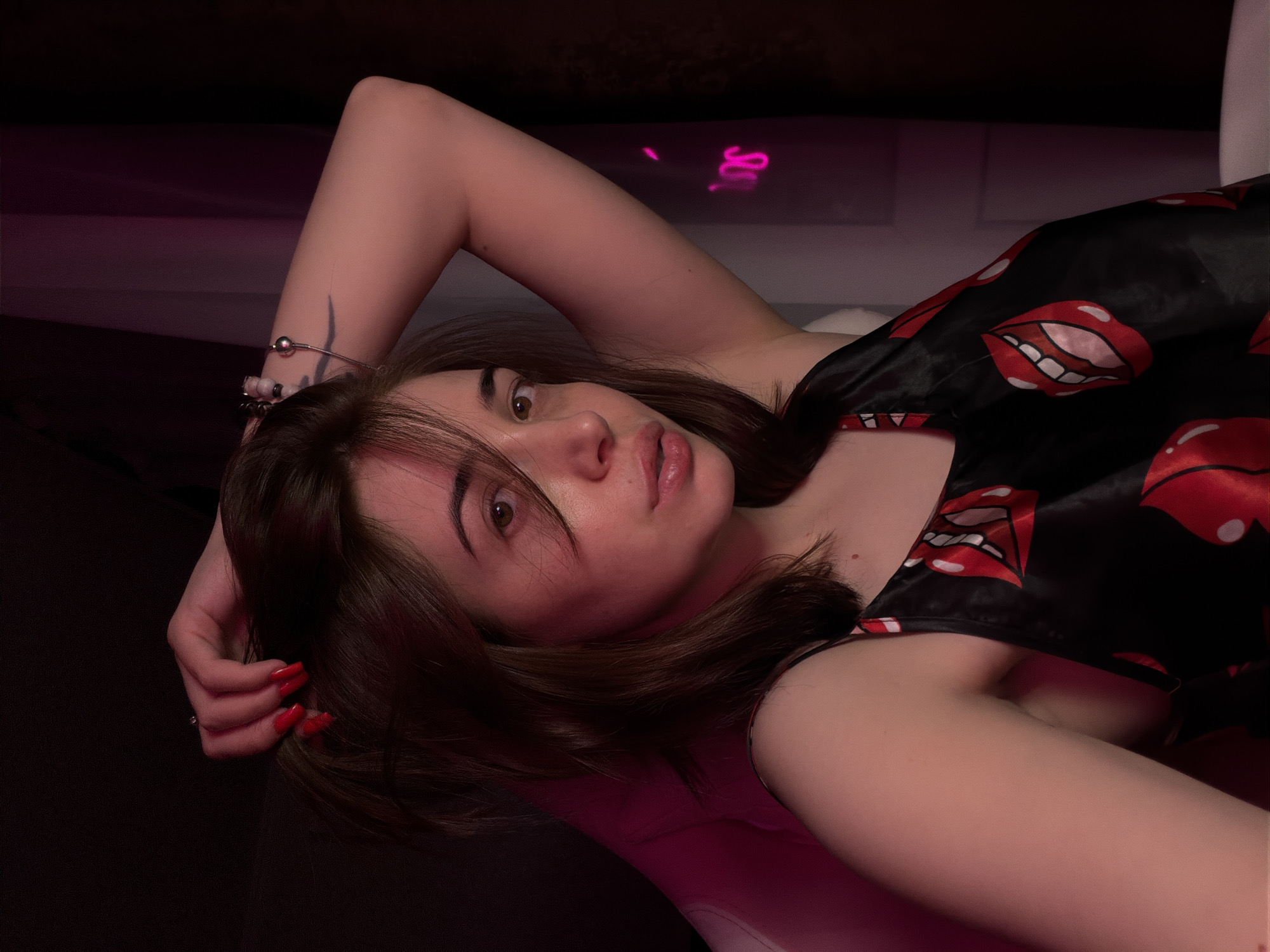 This media file shows a young woman lying down, with her head propped up on her arm. She has brown hair, brown eyes, and fair skin. She is wearing a black satin nightgown with a red lip print pattern. Her fingernails are painted red. On her left wrist, she is wearing a bracelet with silver beads and pink accents. Her right arm is bent, with her hand resting near her head.
The scene appears to be indoors, with dark surroundings and a pinkish glow from what looks like neon lights in the background. There is a blurred neon sign visible in the background, which appears to spell out "SOU". The lighting suggests it might be nighttime or a dimly lit room. The woman's expression is relaxed, and she is looking towards the camera. The overall mood is intimate and casual.