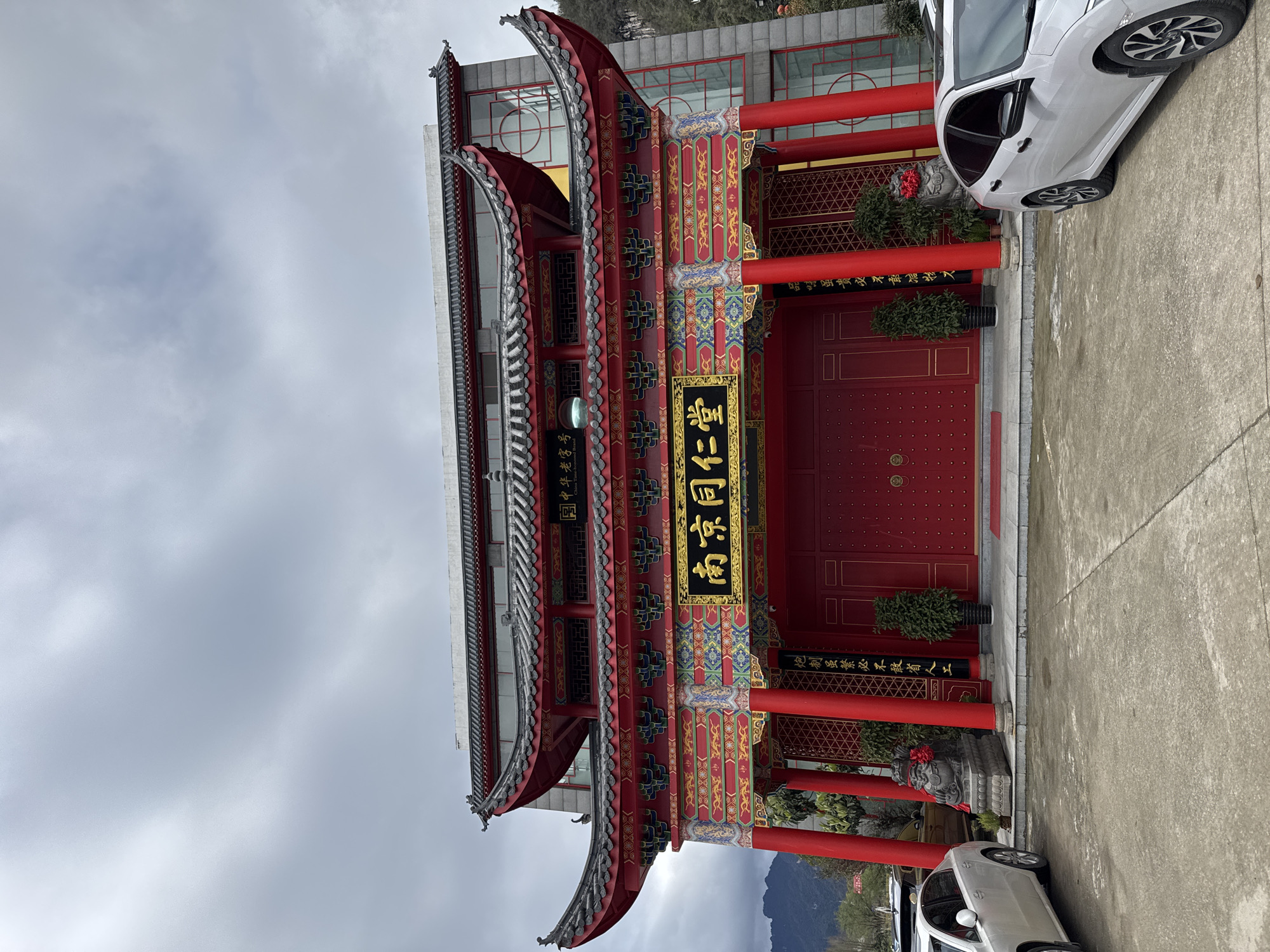 A wide-angle, eye-level, full-frontal shot captures a traditionally styled building facade in Wulingyuan, China, under an overcast sky. The primary structure features a prominent red and gold color scheme with intricate traditional Chinese decorative patterns. A large, horizontal golden plaque centrally positioned above the main entrance displays the Chinese characters "南京同仁堂" (Nanjing Tong Ren Tang). Above this, a smaller sign reads "中華老字號" (China Time-honored Brand). The main entrance consists of large red double doors with golden circular handles, flanked by four substantial red support pillars. Two stone Foo dog statues, each adorned with a red ribbon around its neck, are positioned at the base of the outer pillars. Potted green plants are placed near the entrance. This ornate facade is integrated with a more contemporary building constructed of grey panels and extensive glass windows, visible behind and above the traditional structure. A concrete paved area occupies the foreground, where a white sedan is parked to the left and another white sedan to the right, partially visible. Distant, hazy mountains are discernible on the horizon to the left under the grey sky. No individuals are present in the scene.