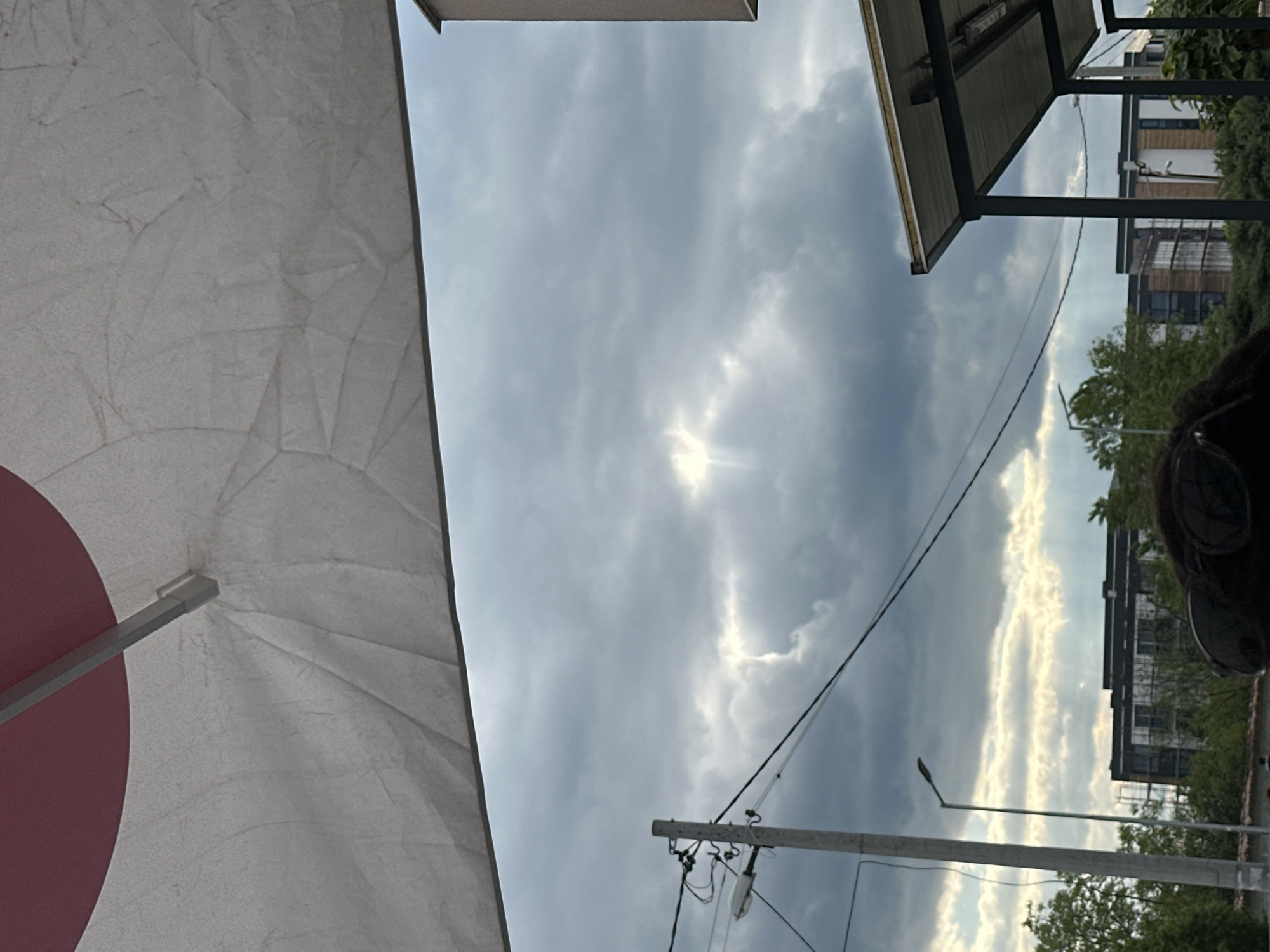 The image, shot from a low angle, showcases a cloudy sky in Plovdiv, Bulgaria. A partial view of a white and red umbrella occupies the left side of the frame. Overhead, a metal structure partially obscures the sky, juxtaposed with buildings peeking through trees in the distance. A utility pole with attached wires and a street light are visible against the backdrop of clouds. An individual with dark hair and sunglasses is also partially in view on the right side.