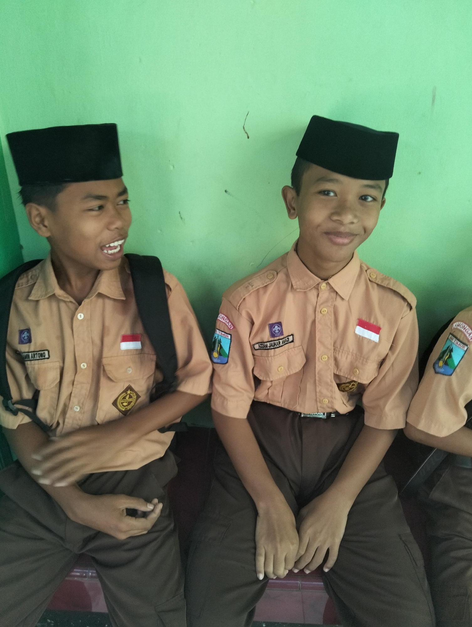 Two young boys, likely students, are pictured sitting next to each other against a light green wall. Both boys are wearing tan uniforms with dark brown pants and black peaked caps, suggesting they might be in uniform for a school or scout activity in Paciran, Indonesia. The boy on the left has his backpack on and appears to be mid-sentence, with his mouth open and teeth visible. The boy on the right is looking towards the camera with a slight smile. Patches are visible on their uniforms, including the Indonesian flag. The wall behind them has some scuff marks and a faint vertical streak. The lighting is even, indicating an indoor or shaded outdoor setting.
