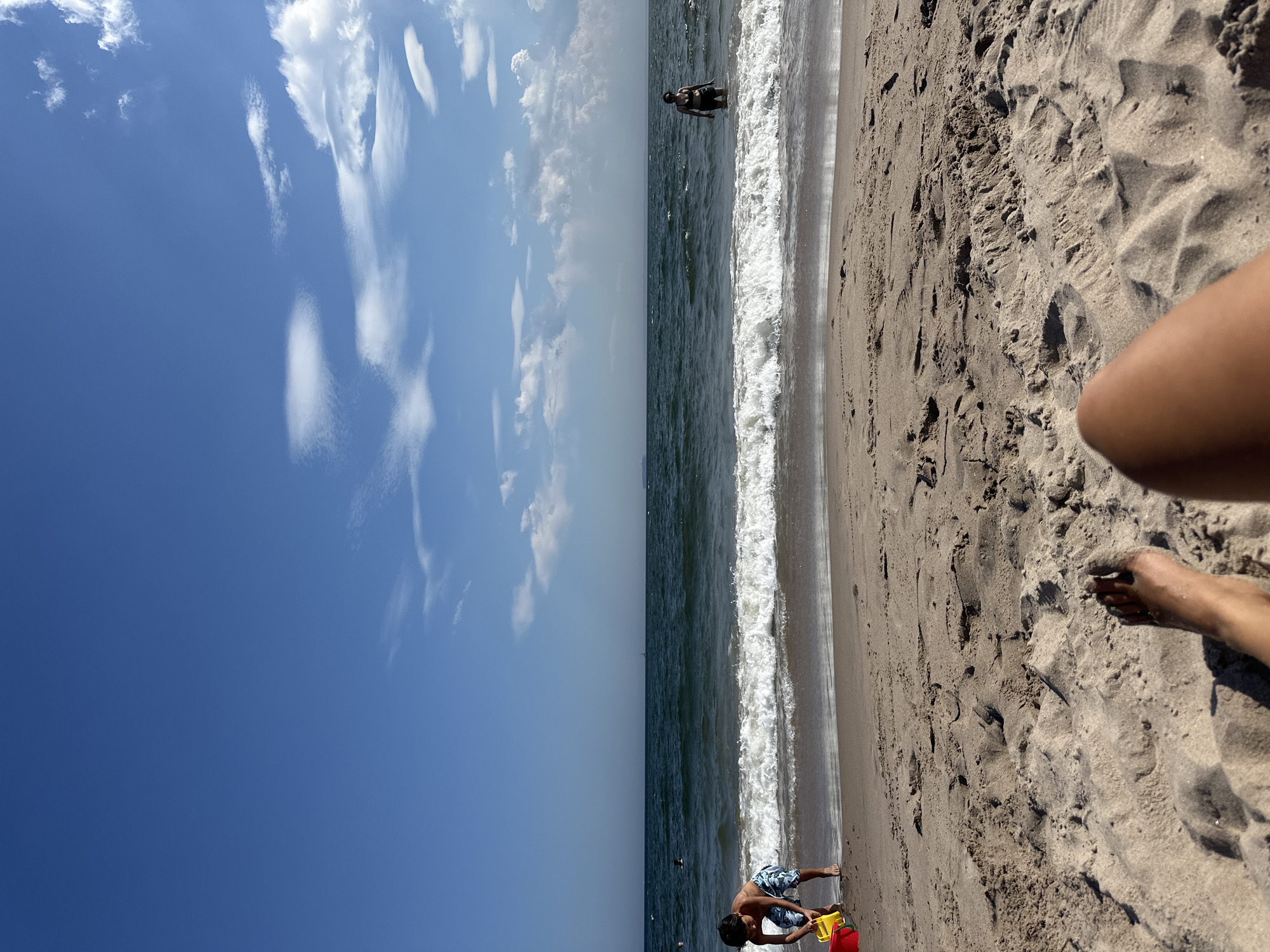 A low-angle shot captures a person's legs and feet buried in the sand on a beach, with the ocean and sky dominating the background. In the middle ground, a child in patterned swim trunks plays in the shallow water with a yellow pail and red bucket. Further out in the ocean, a person in a dark swimsuit stands in the water. The waves are breaking gently on the shore, and the sky is a clear blue with scattered white clouds. The scene appears to be a typical summer day at a beach in New York City, United States of America.