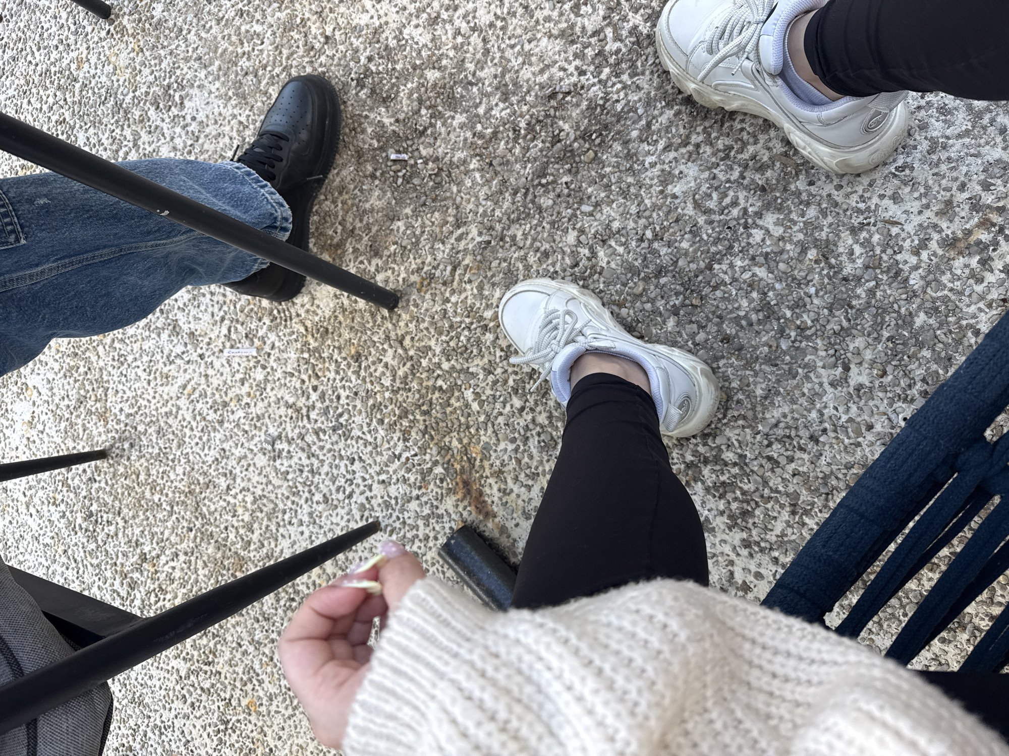 A high-angle shot depicts an outdoor scene with three individuals seated at tables on a textured concrete surface. The person on the left wears blue jeans and black shoes, their legs crossed under the chair. In the middle, another person is wearing black leggings and white sneakers, their chair partially visible. On the right, a third person is wearing black leggings and white sneakers, sitting on a blue chair, their sweater in view. The scene suggests a casual gathering. It is not possible to definitively confirm a location in the city of Varna, Bulgaria, without additional information.