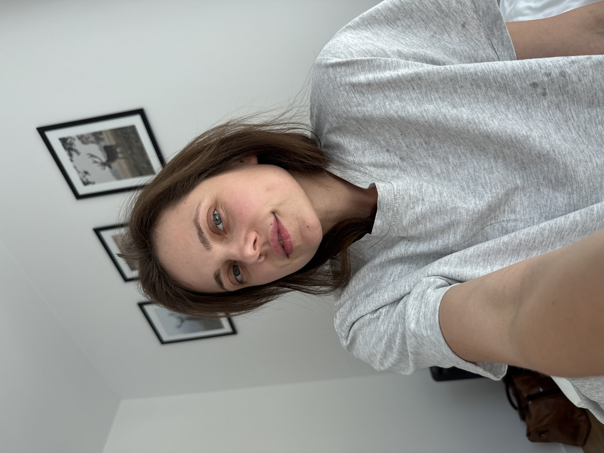 This media file is a selfie taken by a young woman indoors. She is wearing a light gray sweatshirt. Her hair is brown and shoulder-length, and she has green eyes. The woman is looking directly at the camera with a neutral expression. In the background, there are three framed pictures hanging on a white wall. The images in the frames appear to be black and white photographs of nature scenes, possibly including a deer or moose. The lighting suggests it is daytime. The overall setting seems to be a domestic interior, likely a home. There is no visible text in the image.