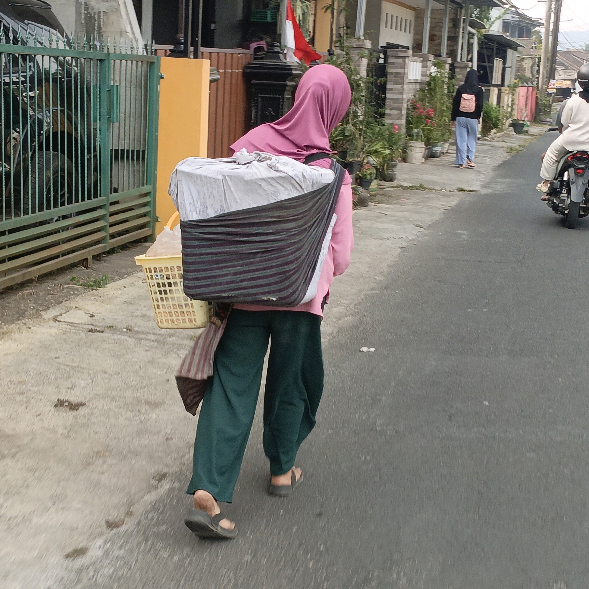 The image depicts a street scene in Salatiga, Indonesia, on an overcast day. A woman wearing a pink hijab and loose green pants is walking away from the camera, carrying a large bundle on her back. The bundle appears to be a woven basket filled with goods, covered by a striped cloth and a white sheet. She is wearing dark sandals. To her left, a green metal gate with vertical bars encloses what appears to be a yard with parked motorcycles. A bright yellow pillar stands beside the gate. In the background, residential buildings with tiled roofs line the street, interspersed with plants in pots and some small trees. Further down the road, another person in a black hoodie and light blue pants is walking away from the camera, and a motorcycle with a rider wearing a white top is visible on the right side of the frame. The road surface is asphalt.