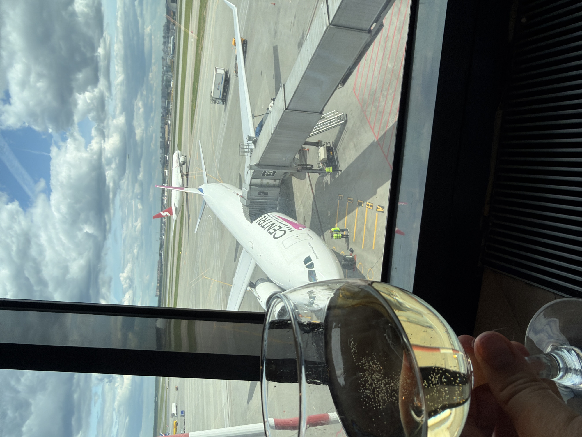 The image shows a view from a window at an airport. Visible through the window is an airplane parked at a gate, connected to a jet bridge. Airport workers are visible near the plane. In the foreground, a hand holds a glass of champagne or sparkling wine. The reflection in the window shows cloudy skies and distant buildings. Based on the text on the plane saying "CENTRUM Air", the location is Khimki, Russia.