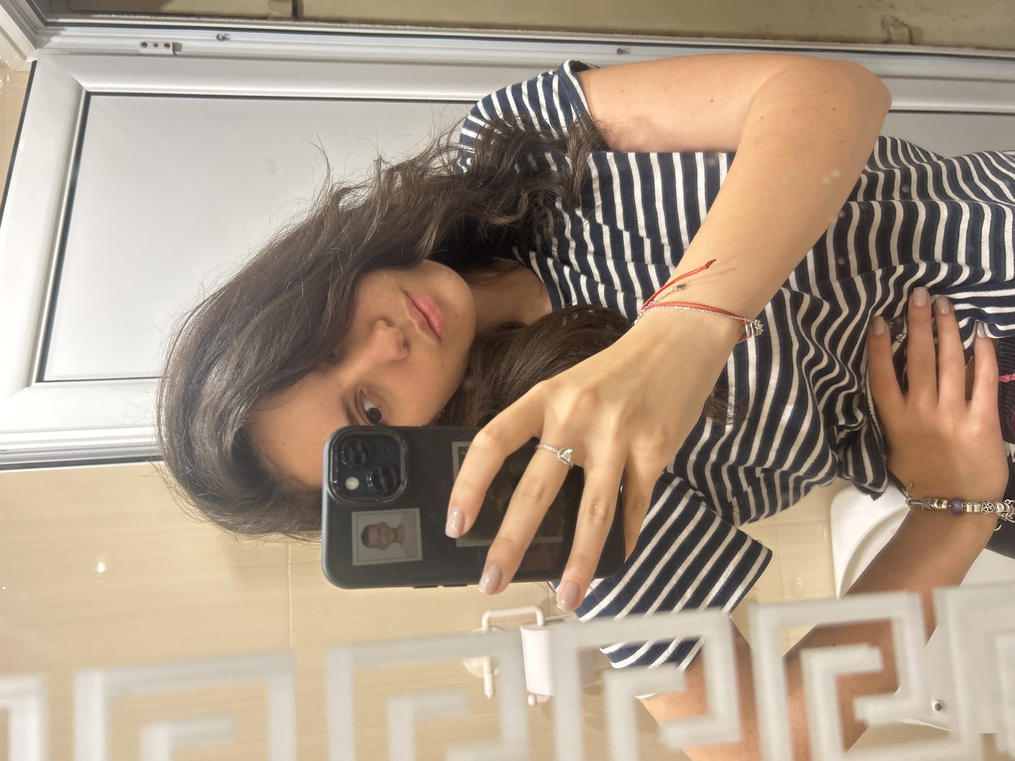 A woman with dark hair is taking a selfie in a bathroom. She's wearing a striped navy and white shirt. She has a ring on her finger and bracelets on her wrist. Her phone is black, and a photo of a person is visible on the back of the phone. The bathroom has beige tile walls, a white trim, and a white towel holder. The location is Unknown, Unknown.